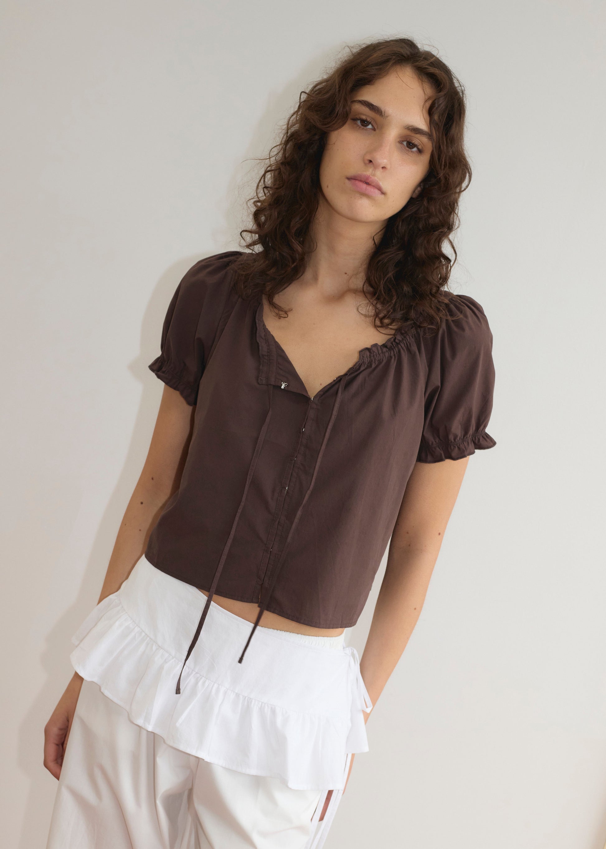 A young woman with wavy brown hair wears a brown short-sleeve blouse and the Flounce Belt - White, which adds a lightweight frill ruffle detail to her pants. She stands against a plain off-white background, looking at the camera.