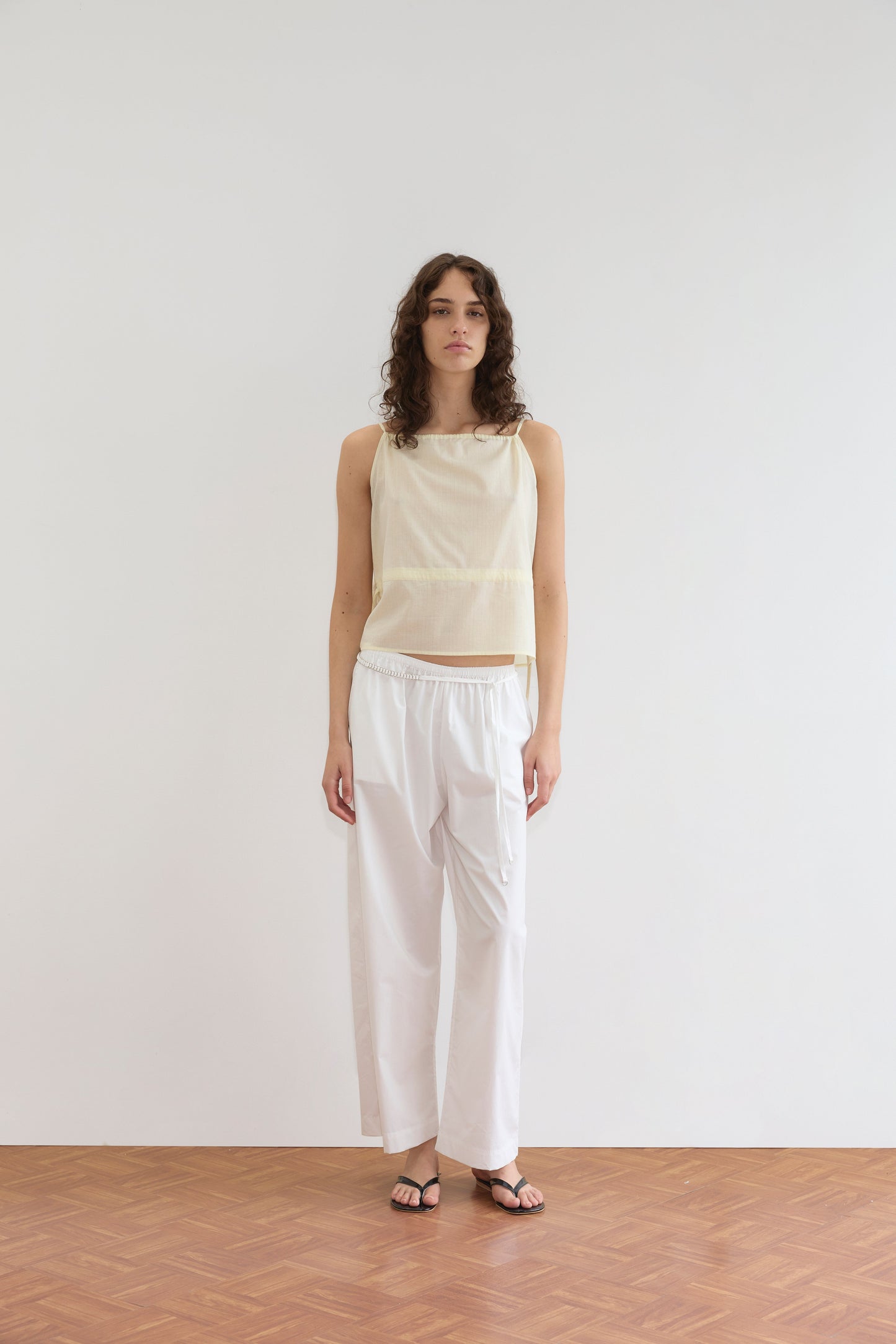 A person with long curly hair wears a Deiji Studios Waist Tie Top in soft yellow stripe, paired with loose white pants and black sandals, standing against a plain white wall on a wooden floor.