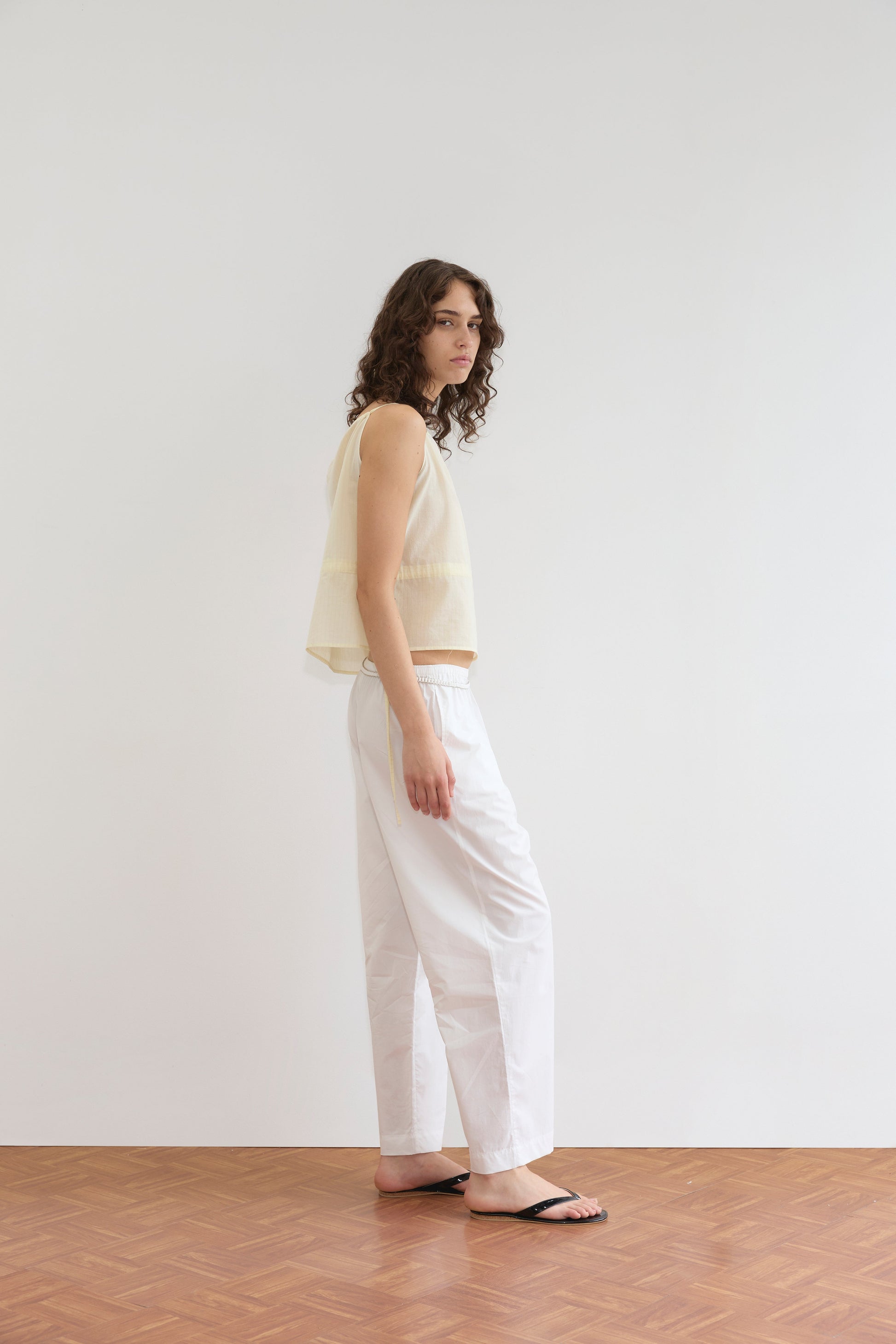 A person with long curly hair stands sideways on a wooden floor, wearing the Deiji Studios Waist Tie Top in Soft Yellow Stripe, loose white pants, and black flip-flops, against a plain white wall.