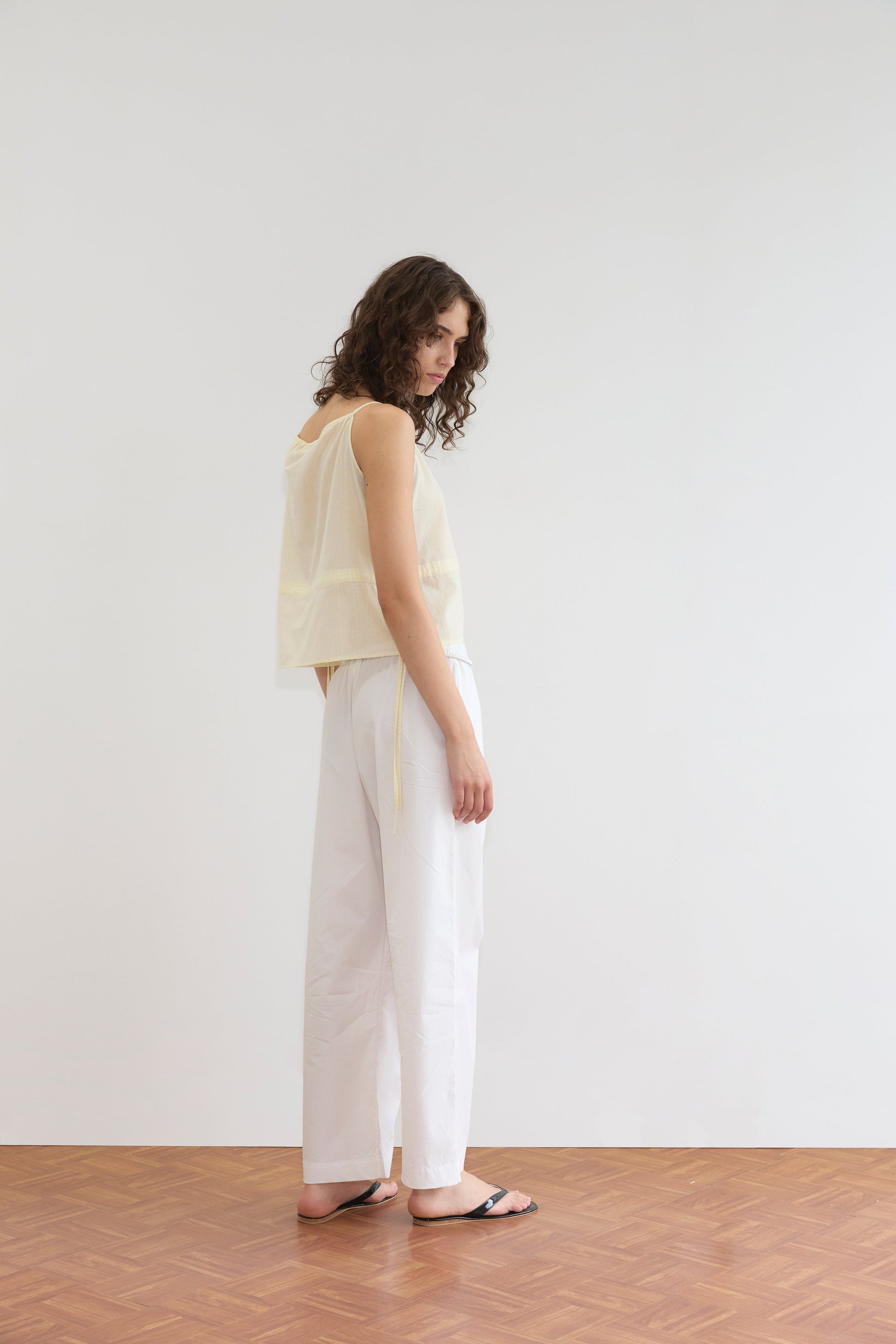 A person with curly hair wears the Deiji Studios Waist Tie Top in Soft Yellow Stripe, paired with loose white pants and black sandals, standing on a wooden floor against a plain white background while looking over their shoulder.
