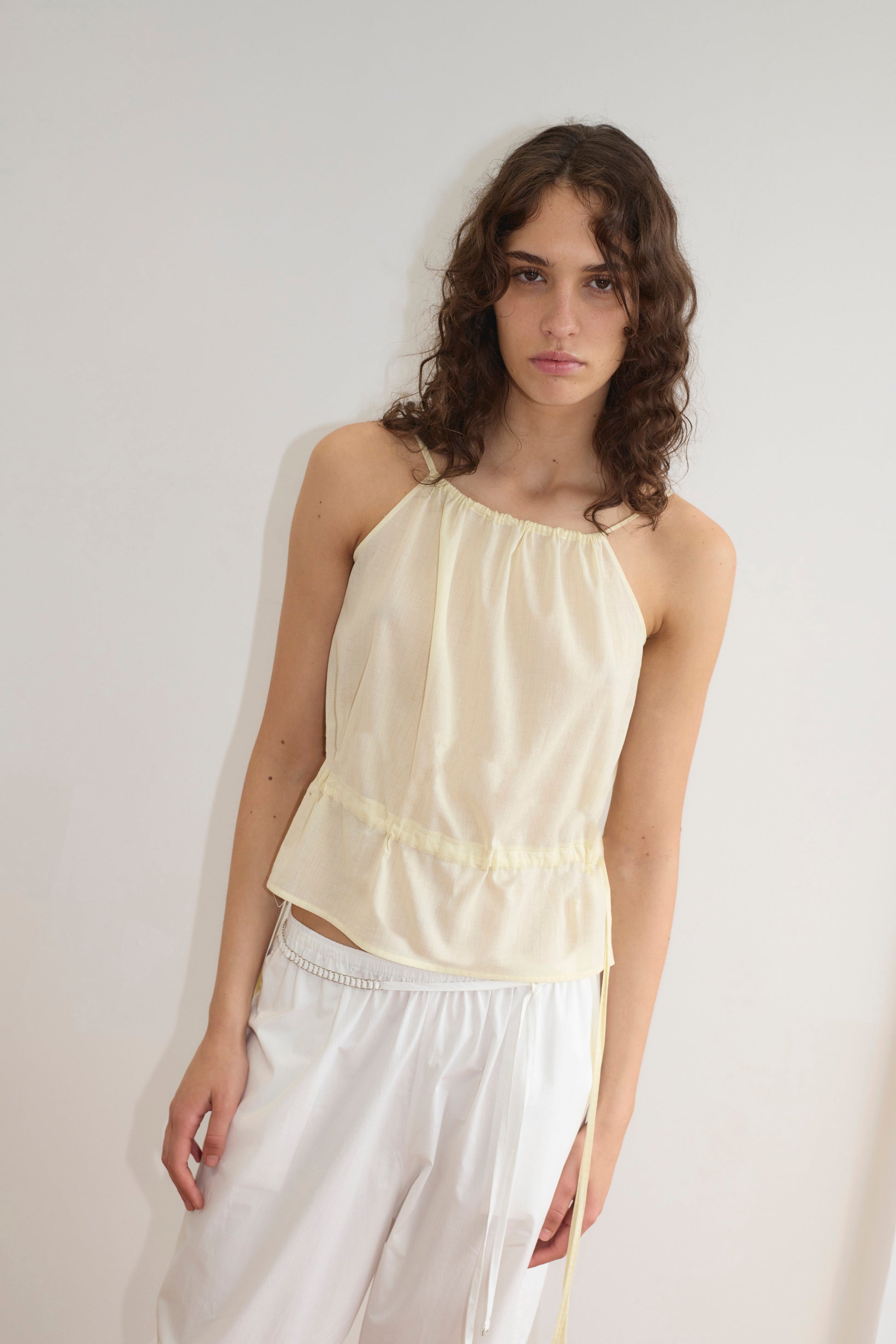 A young woman with wavy brown hair wears the Deiji Studios Waist Tie Top in Soft Yellow Stripe with white pants, standing against a plain white wall and looking at the camera with a neutral expression.