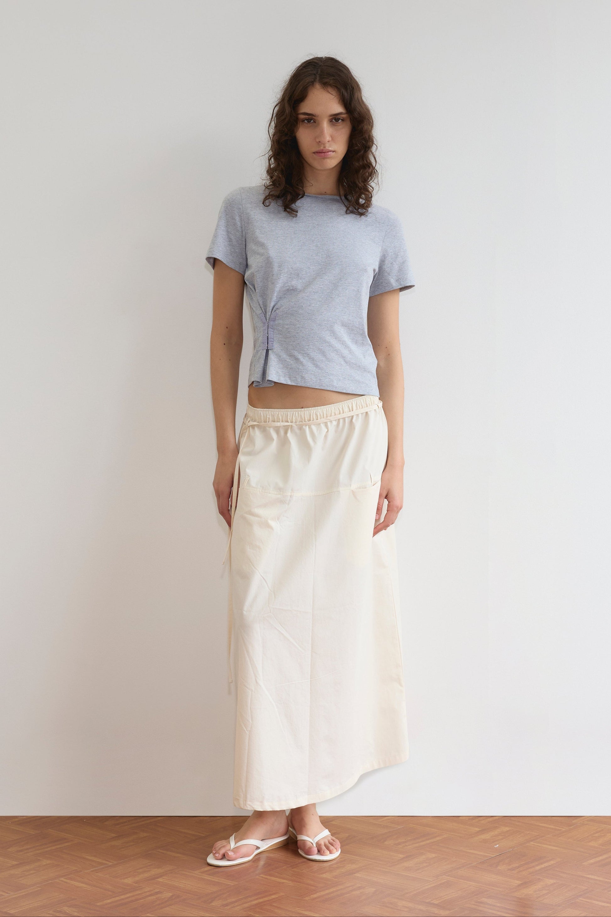 A woman with wavy brown hair and a neutral expression stands against a white wall, wearing the Loop Pocket Skirt - Off White by Deiji Studios, styled with a light purple short-sleeve top and white flip-flops for an effortlessly chic look.