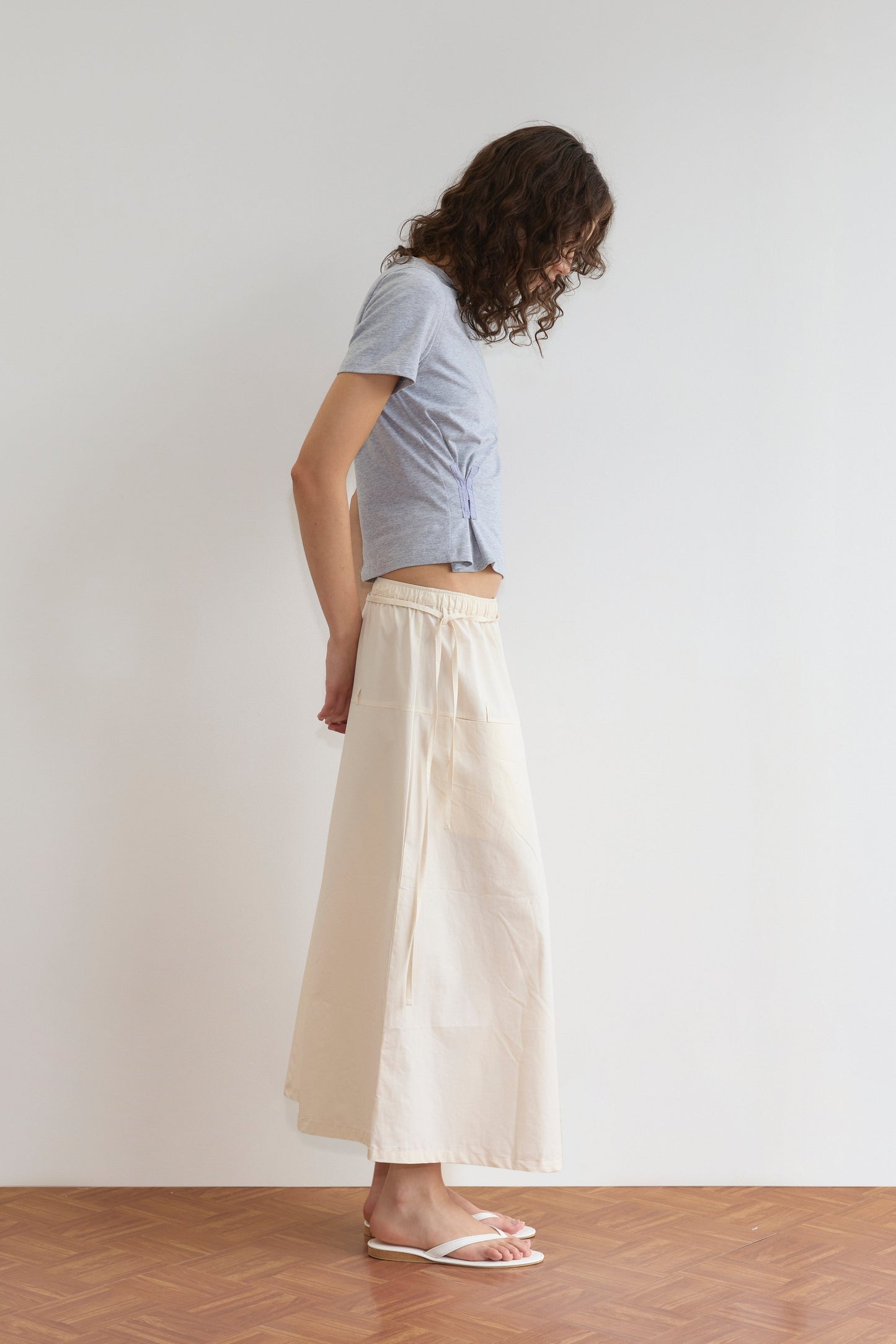 A person with wavy hair stands in profile against a white wall, wearing a light gray t-shirt, the Loop Pocket Skirt - Off White, and white sandals on a wooden floor.