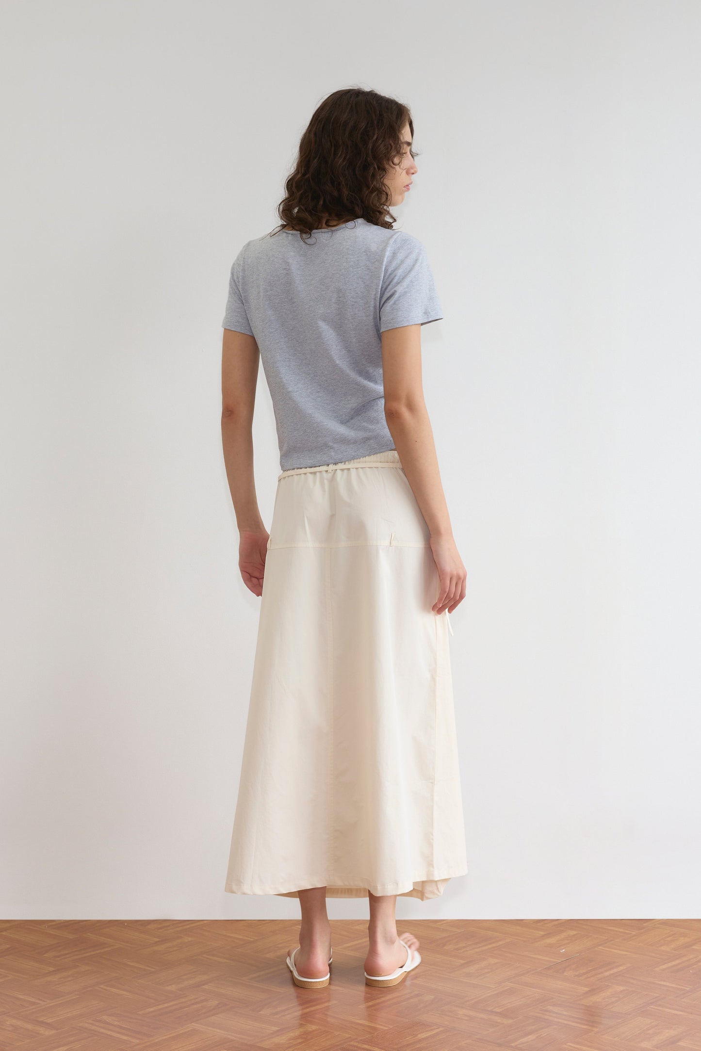 A woman stands indoors with her back to the camera, wearing a light gray short-sleeve t-shirt, the Loop Pocket Skirt - Off White by Deiji Studios, and white sandals on a wooden floor against a plain white wall.