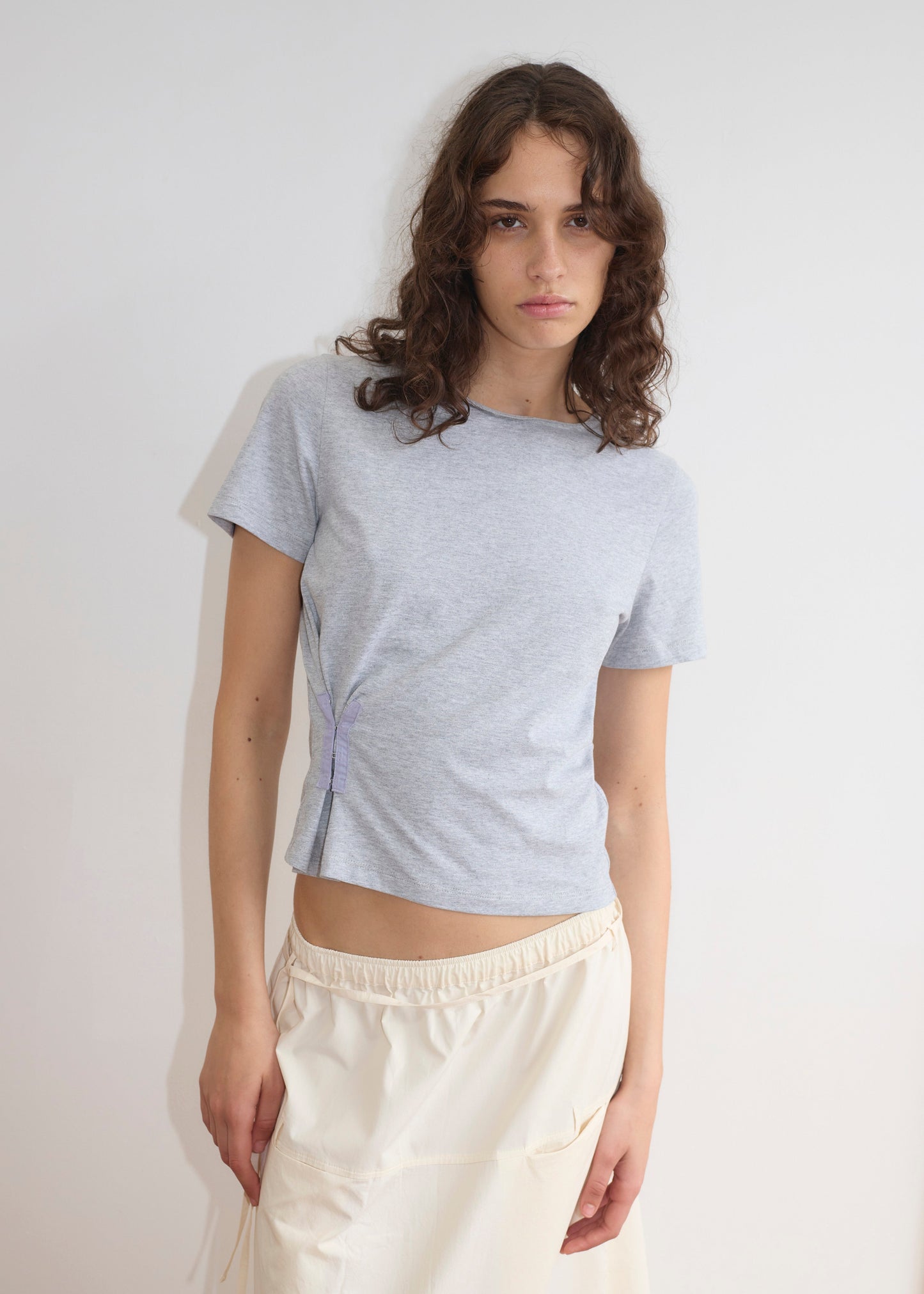 A young woman with wavy brown hair wears a light grey short-sleeve top and the Loop Pocket Skirt - Off White by Deiji Studios. She stands against a plain white background, looking slightly to the side with a neutral expression.