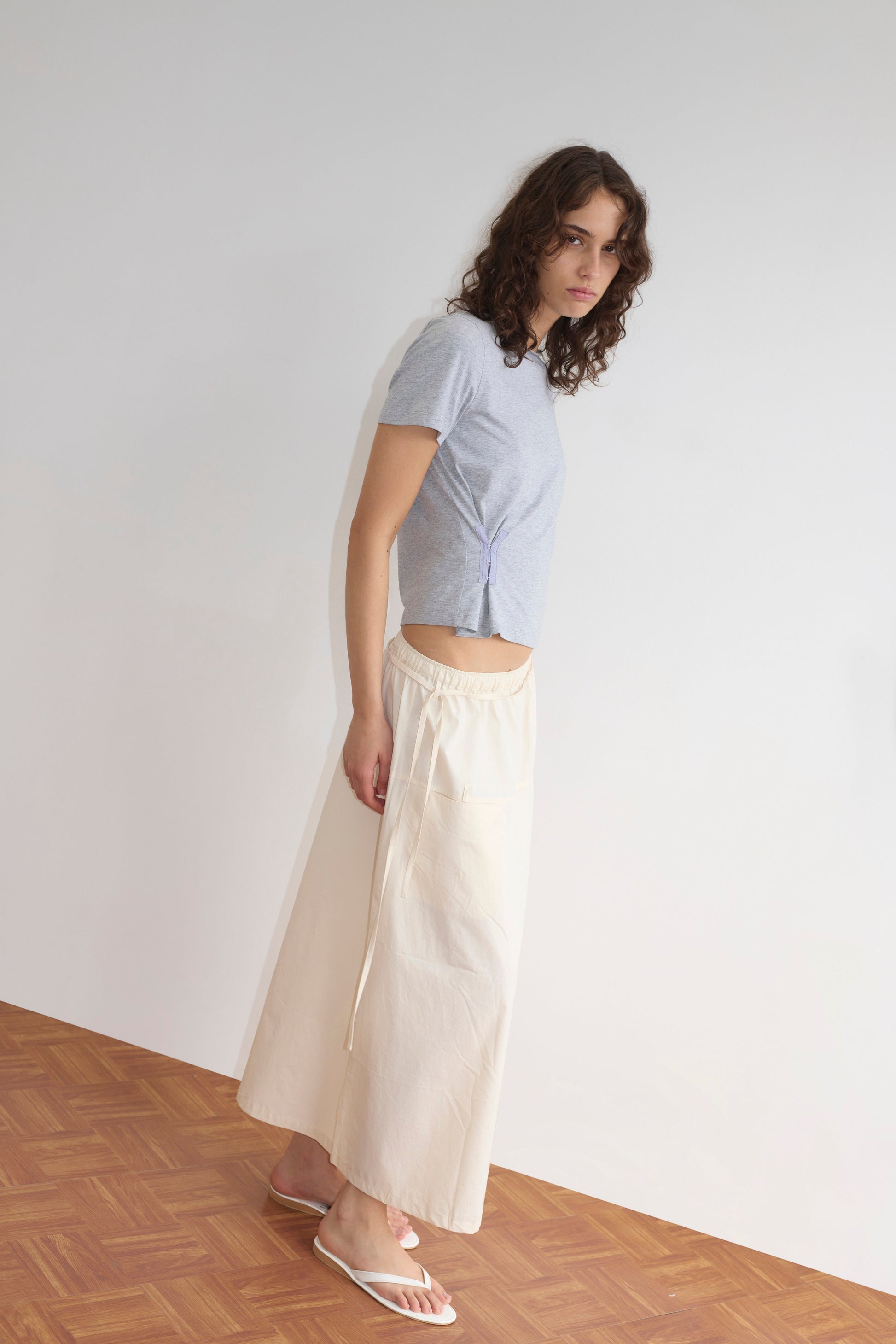 A woman with wavy brown hair stands indoors against a white wall, wearing a light grey short-sleeve top, the Loop Pocket Skirt - Off White by Deiji Studios, and white sandals on a wooden floor.