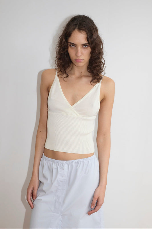 A young woman with wavy brown hair stands before a plain white wall, wearing the Cross Panel Knit Top—a white sleeveless style with thin straps—paired with a light blue skirt. She has a neutral expression and relaxed arms at her sides.