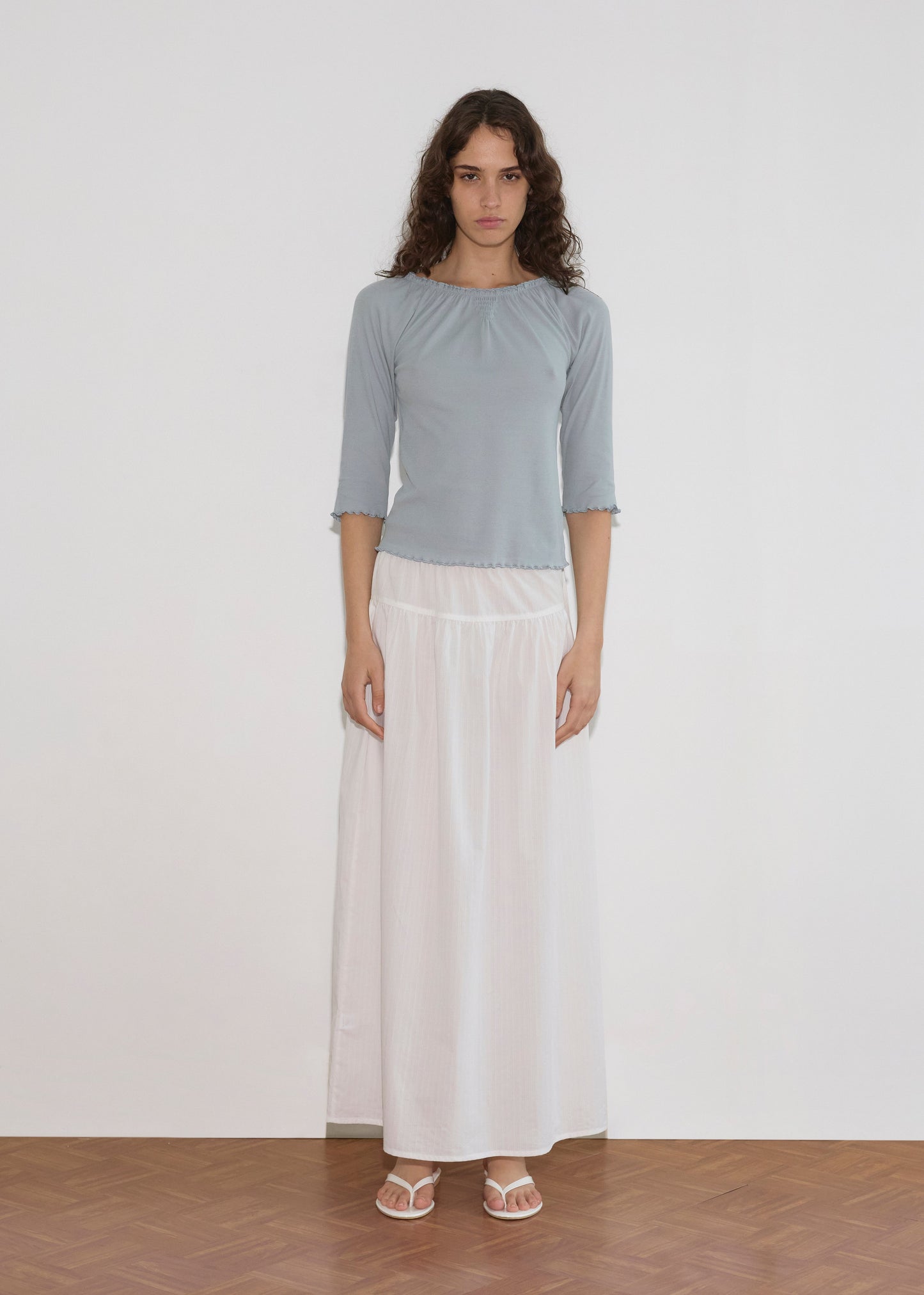 A woman with long curly hair stands indoors against a white background, wearing a light blue top, the Maxi Skirt - White Texture by Deiji Studios, and white sandals. She looks directly at the camera with a neutral expression.