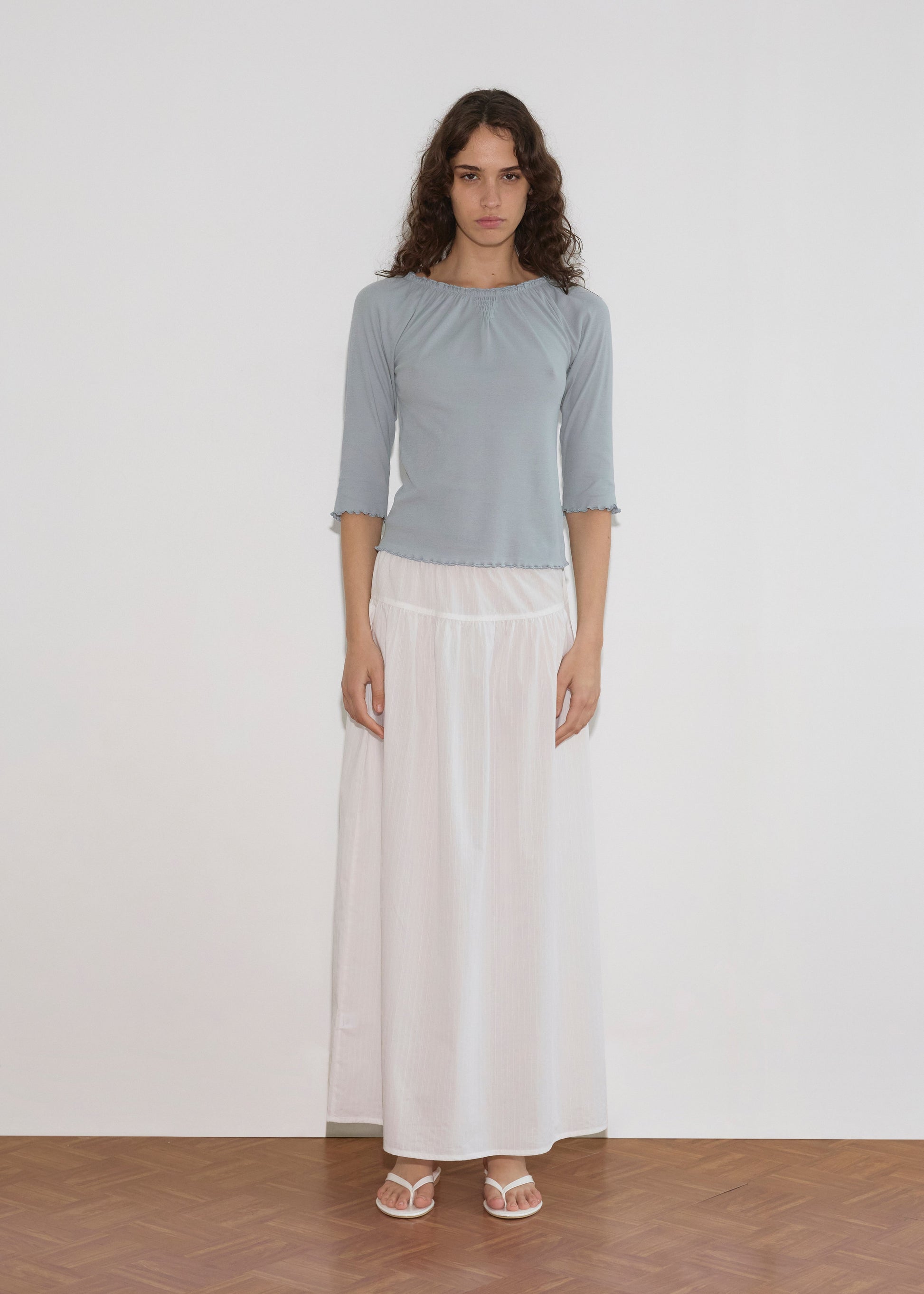 A woman with long curly hair stands indoors against a white background, wearing a light blue top, the Maxi Skirt - White Texture by Deiji Studios, and white sandals. She looks directly at the camera with a neutral expression.