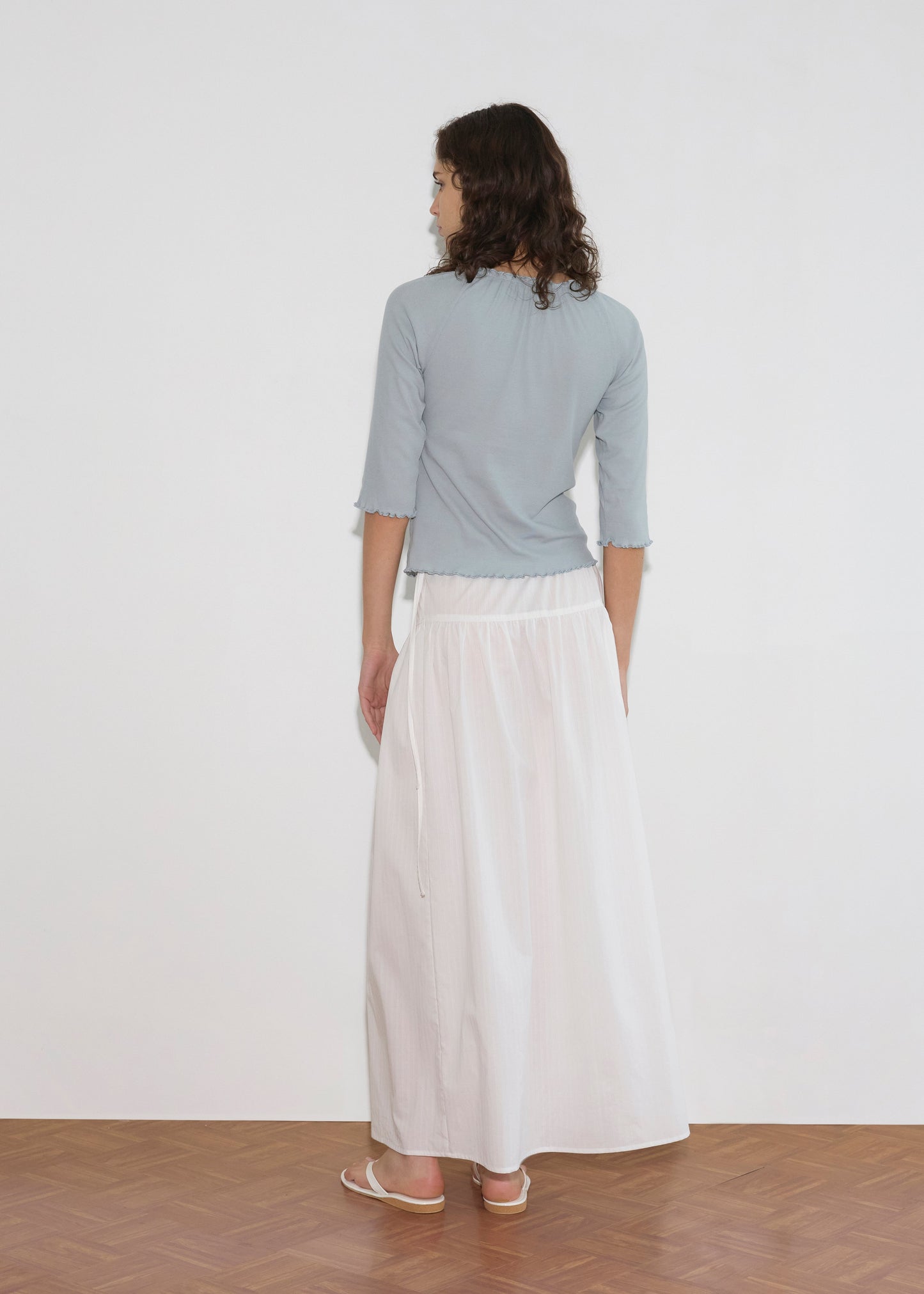 A person with shoulder-length curled hair faces away, wearing a light blue three-quarter sleeve top and the Maxi Skirt - White Texture with white sandals, standing on a wooden floor against a plain white wall.