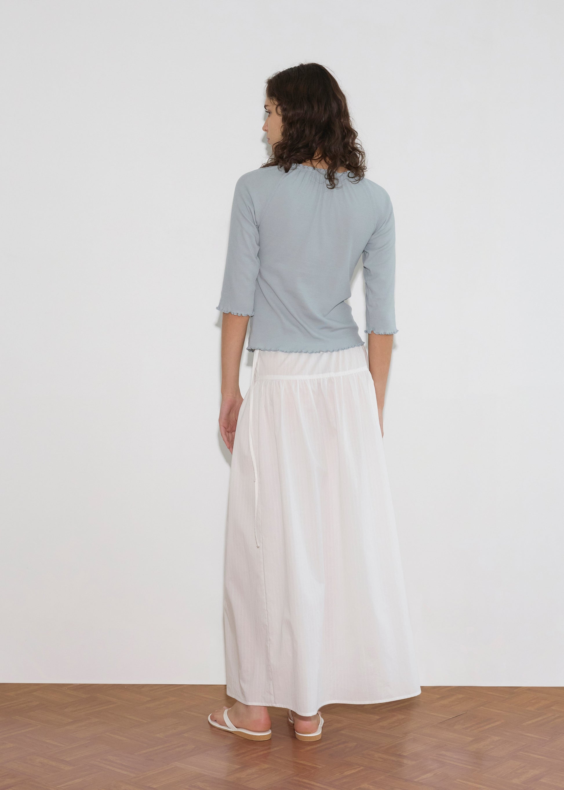 A person with shoulder-length curled hair faces away, wearing a light blue three-quarter sleeve top and the Maxi Skirt - White Texture with white sandals, standing on a wooden floor against a plain white wall.