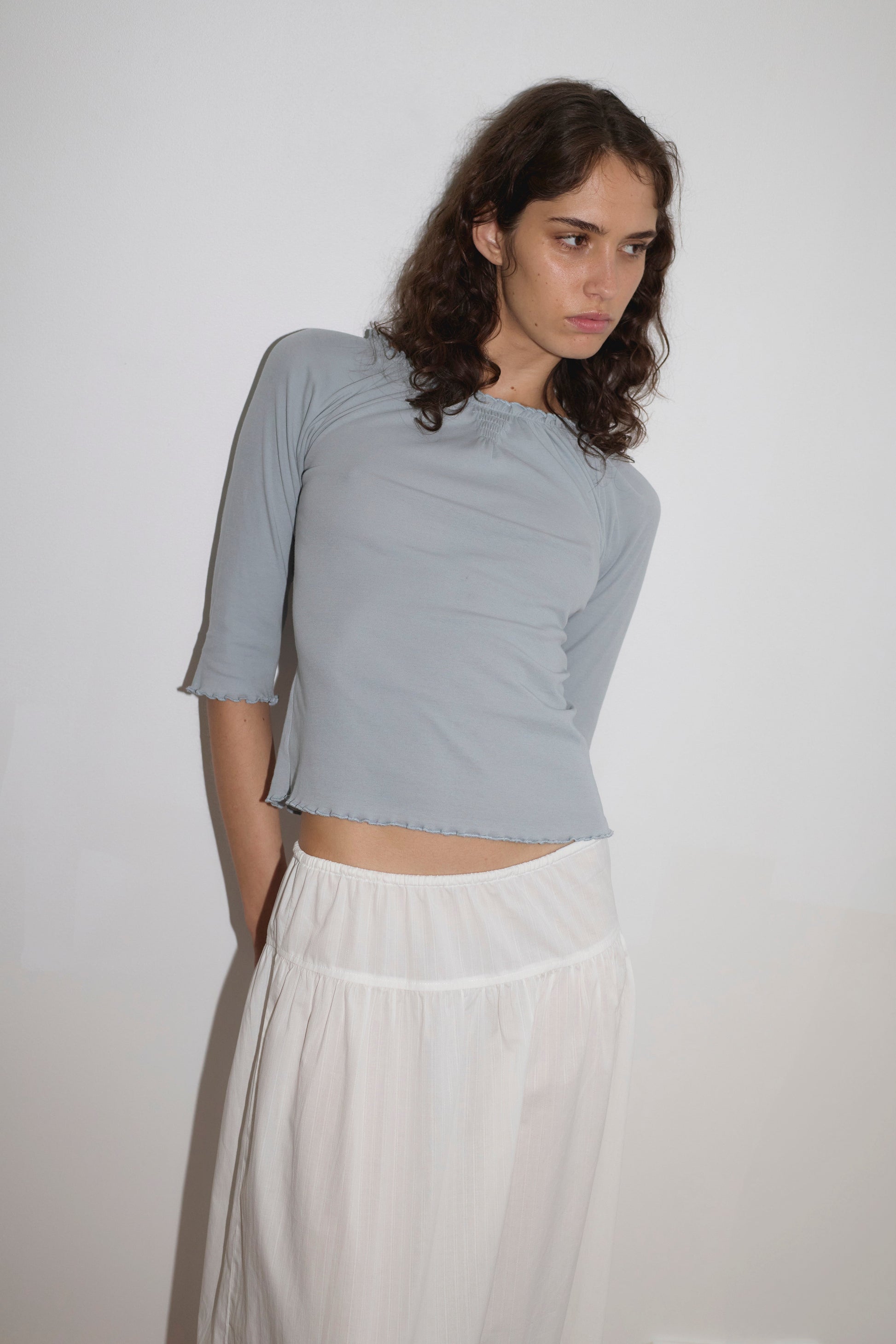 A woman with wavy brown hair in a light blue three-quarter sleeve top and the Maxi Skirt - White Texture stands against a plain white wall, looking to the side with a neutral expression.