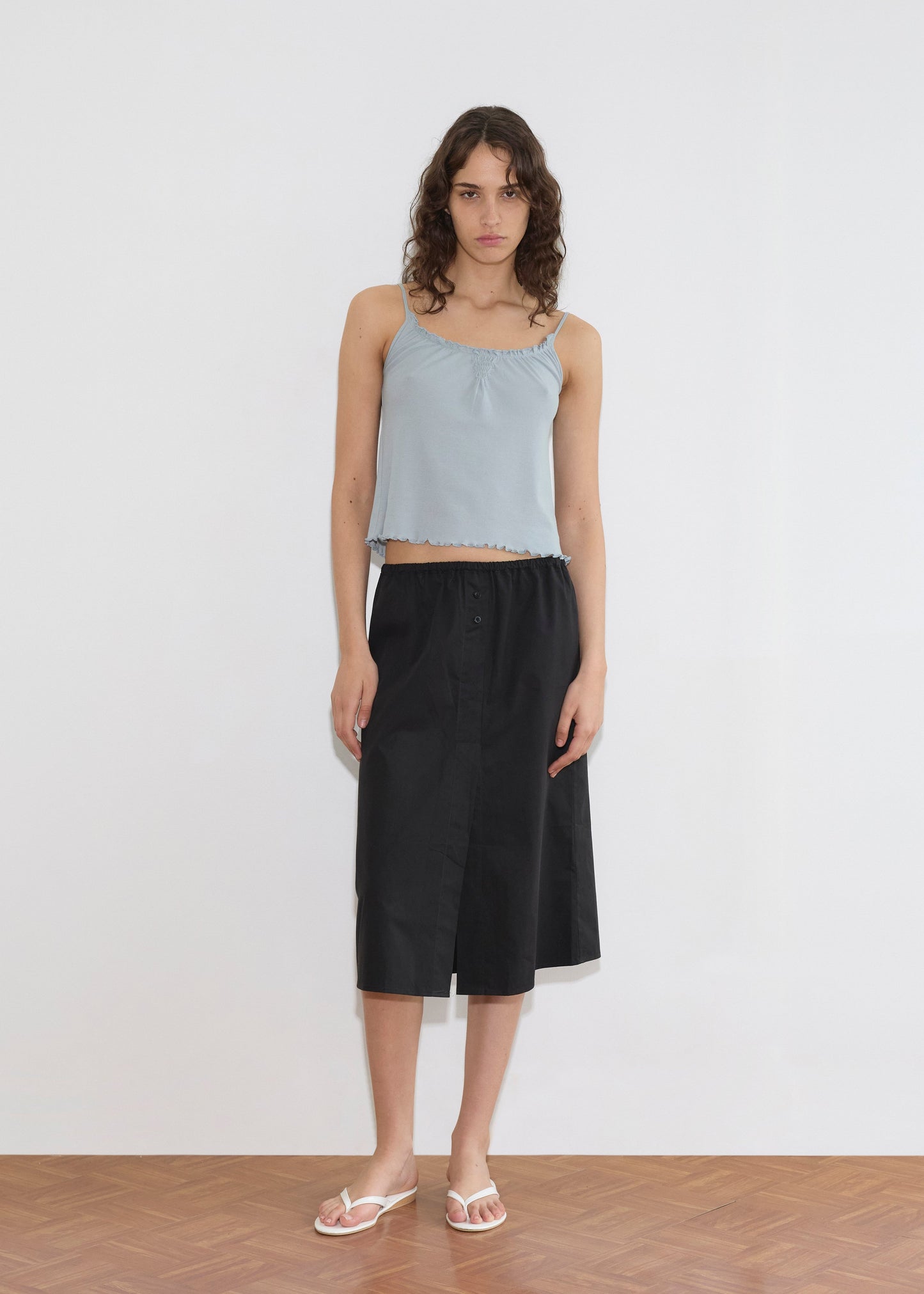 A model wearing a light blue cropped camisole with ruffled trim and the Overlap Skirt—a black midi skirt with an elasticated waist and front slit—paired with white flip-flops, stands against a white wall.