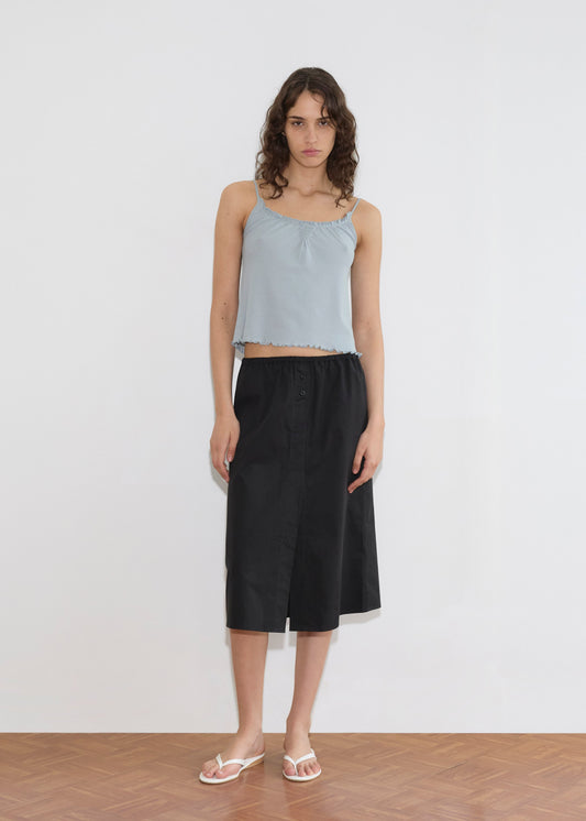 A model wearing a light blue cropped camisole with ruffled trim and the Overlap Skirt—a black midi skirt with an elasticated waist and front slit—paired with white flip-flops, stands against a white wall.