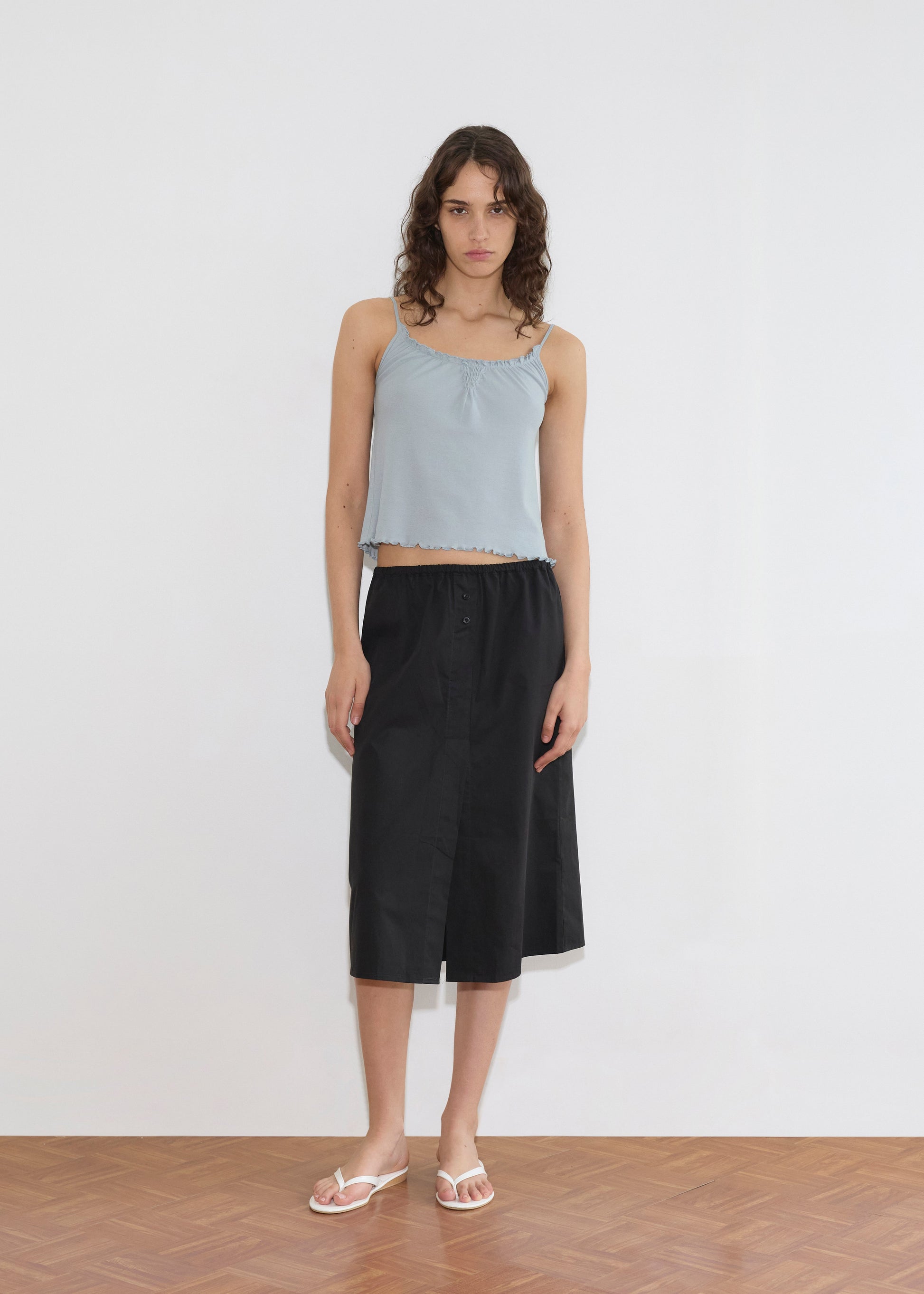 A woman with wavy brown hair wears the Cotton Flounce Tank - Baby Blue, paired with a black knee-length skirt and white flip-flops. She stands on a wooden floor against a plain white wall.