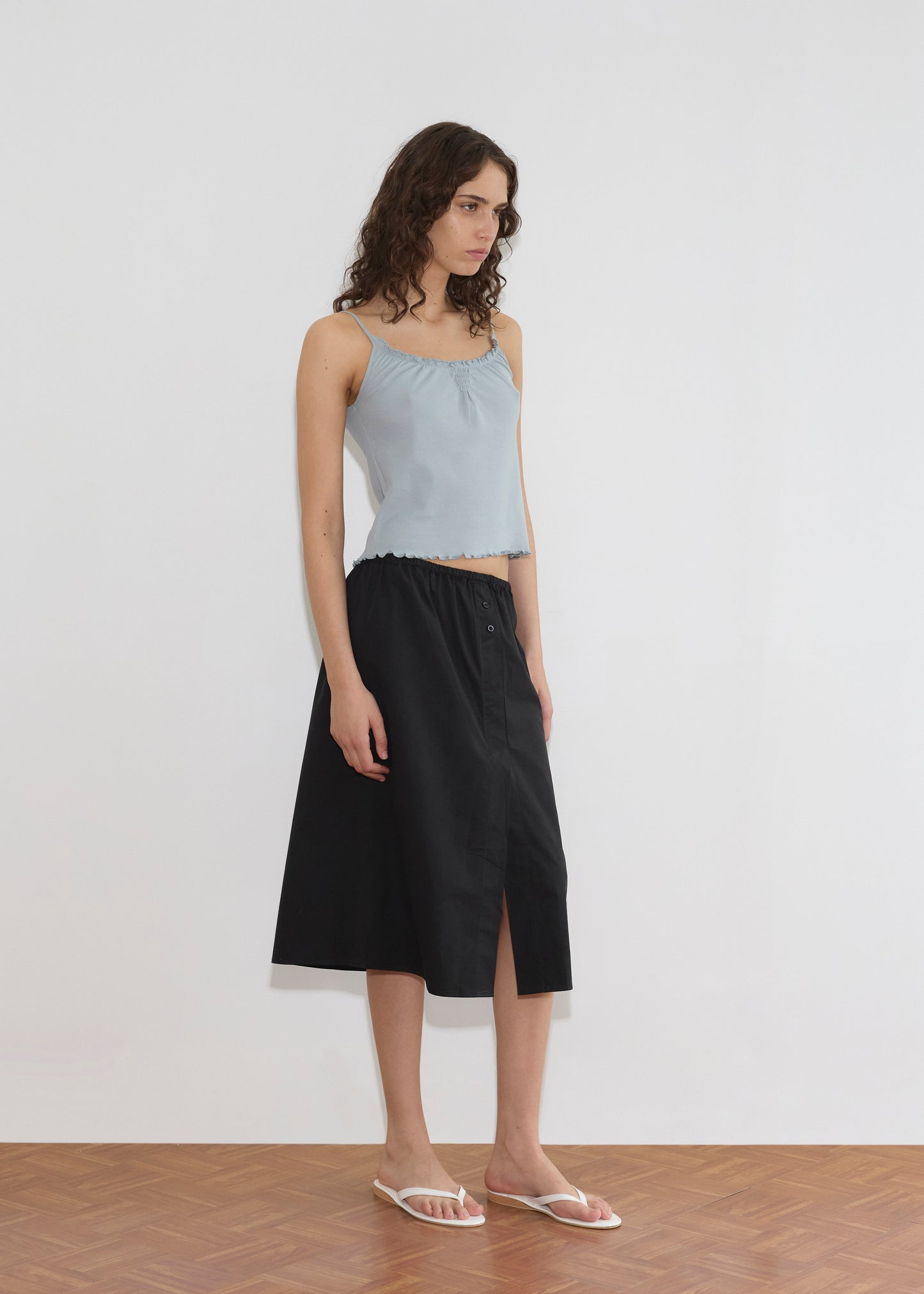 A woman with wavy brown hair wears the Overlap Skirt - Black, featuring an elastic waistband and front slit, paired with a light blue sleeveless top and white flip-flops. She stands indoors against a plain white wall, looking slightly downward.