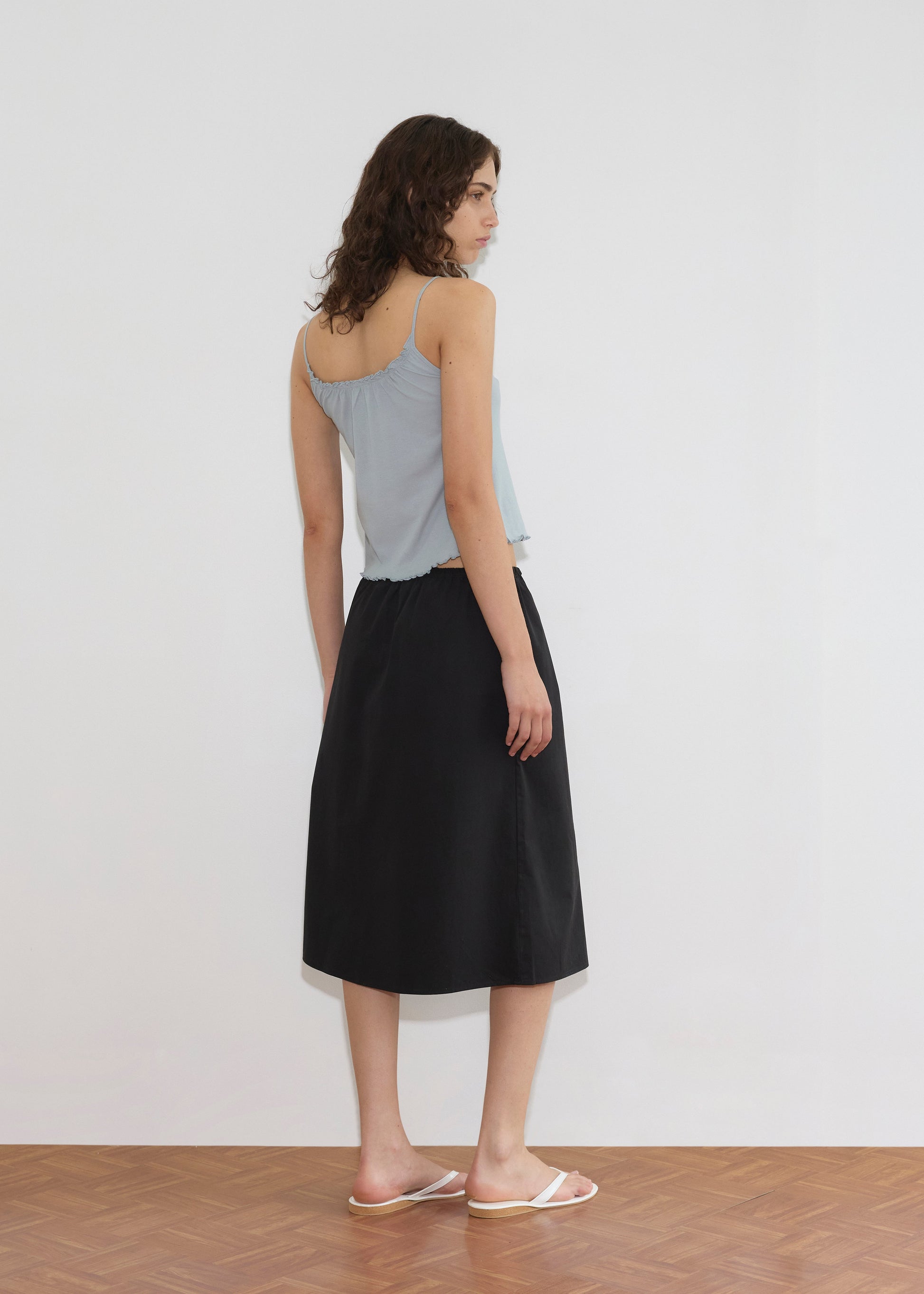 A woman with wavy brown hair stands facing away, wearing the Cotton Flounce Tank - Baby Blue, a black midi skirt, and white slide sandals on a wooden floor against a plain white wall.