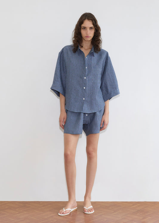 A person with long wavy hair wears the 03 Set - Azure Stripe by Deiji Studios—loose linen shirt and matching boxer shorts—standing barefoot in white flip-flops on a wooden floor against a plain white wall.