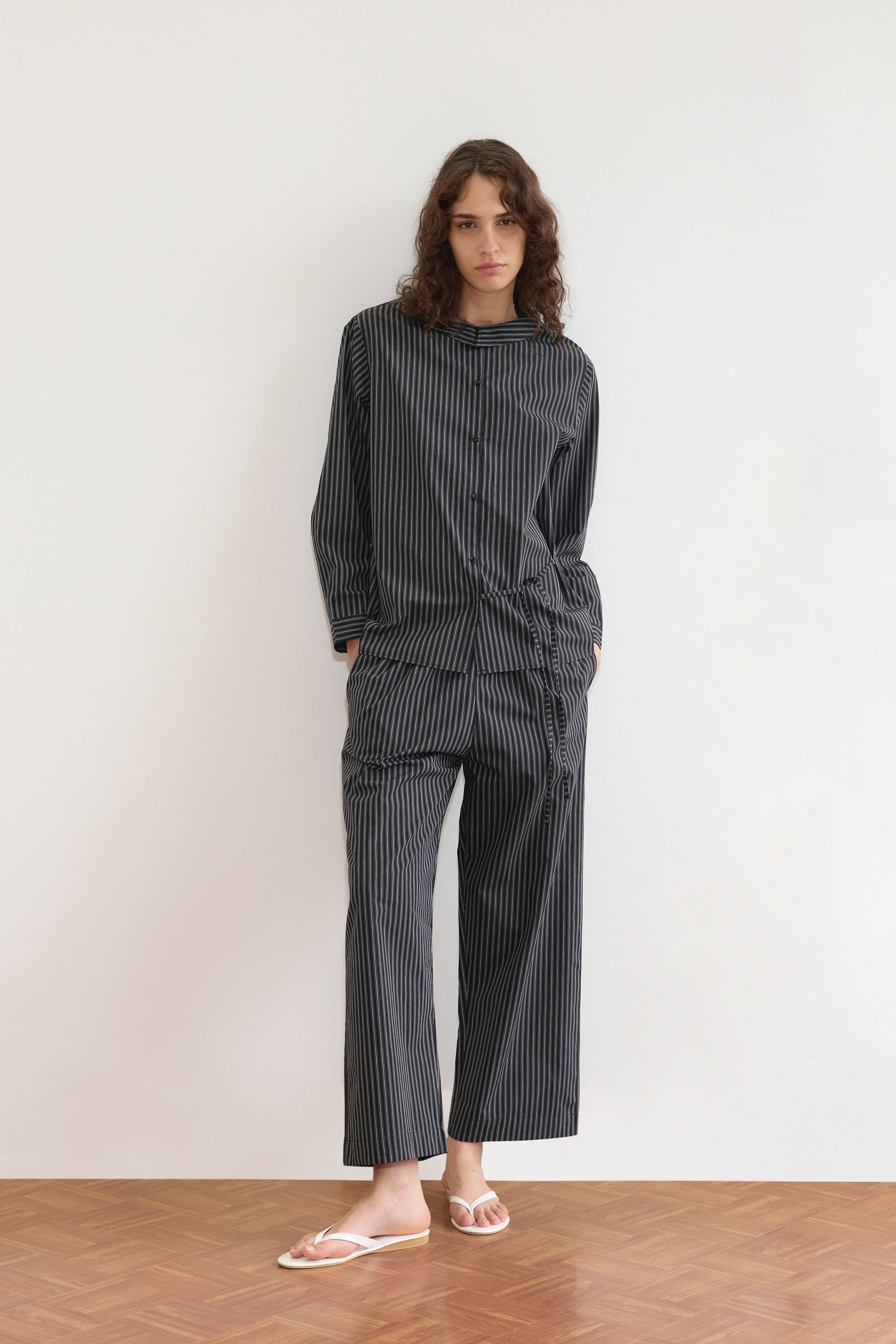 A person with long curly hair stands against a white wall, wearing the Deiji Studios Left Set - Mono Stripe: a lightweight, relaxed shirt and matching pants with hands in pockets, paired with white flip-flops on a wooden floor.