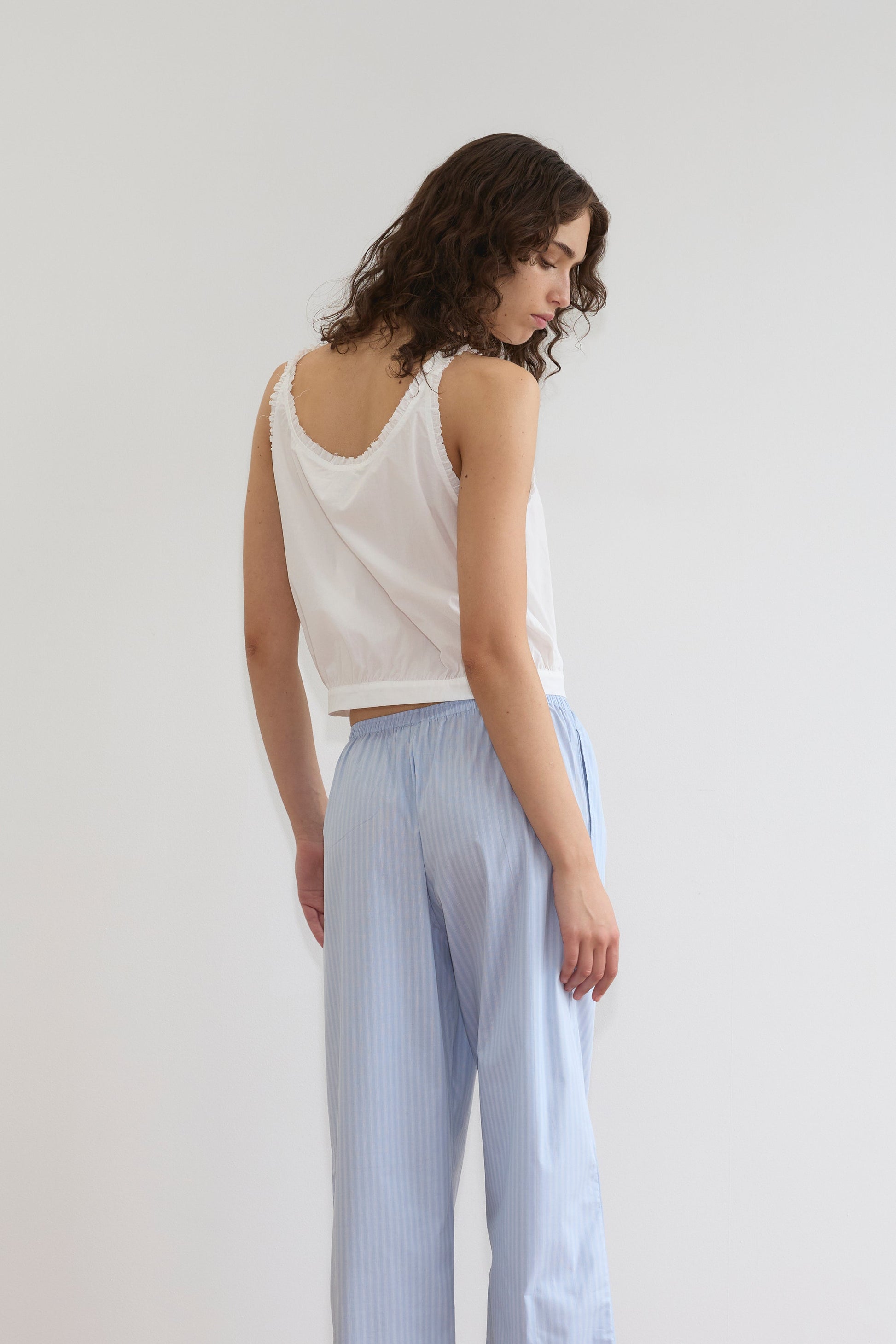 A person with wavy brown hair stands sideways, looking down, wearing the Overlie Top in white and light blue striped pants with a fitted waist, set against a plain white background.