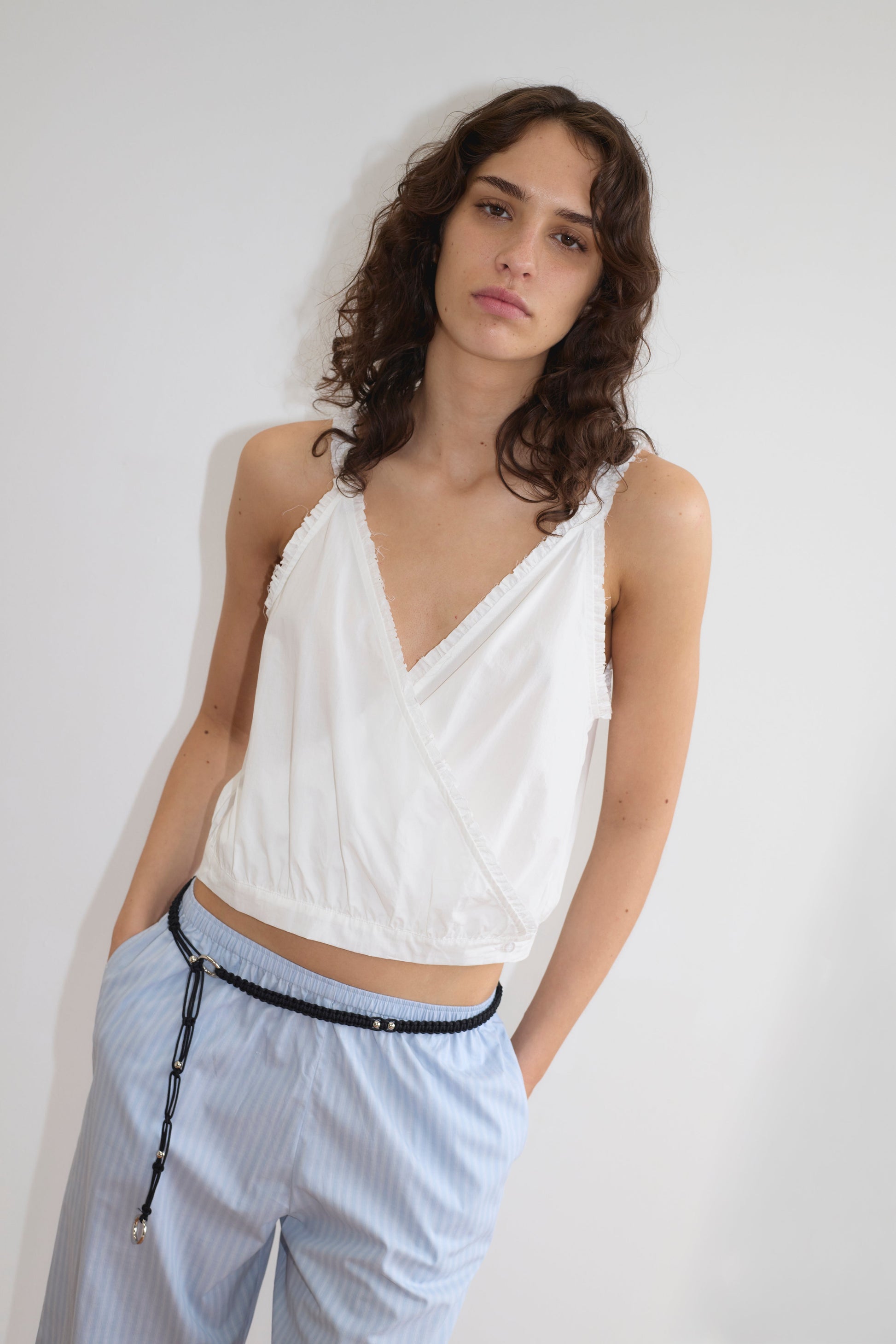 A person with wavy brown hair wears the Overlie Top - White, a sleeveless crossover front wrap top, paired with light blue striped pants with a fitted waist, standing against a plain white background with one hand in their pocket.