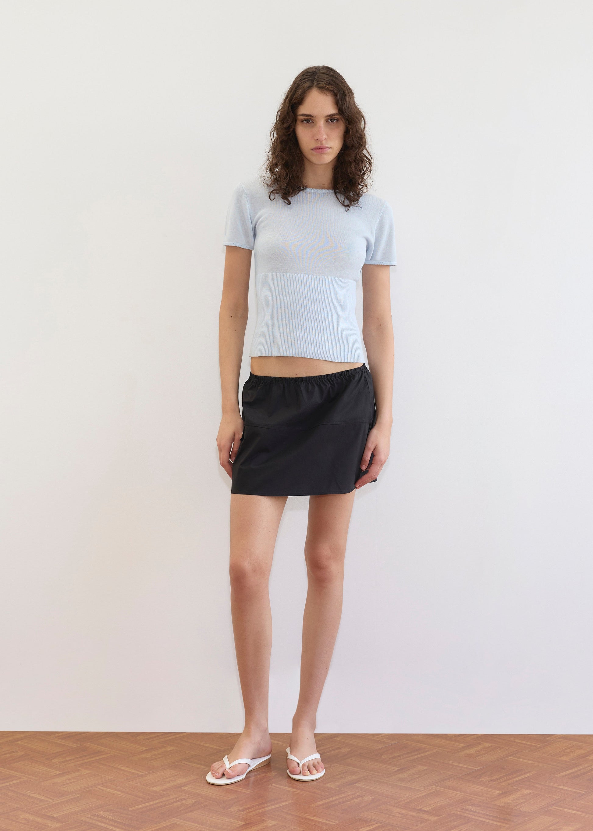 A person with wavy brown hair stands indoors against a white wall, wearing a light blue short-sleeve top, the Pocket Skirt - Black at mini-length, and white sandals on a wooden floor.