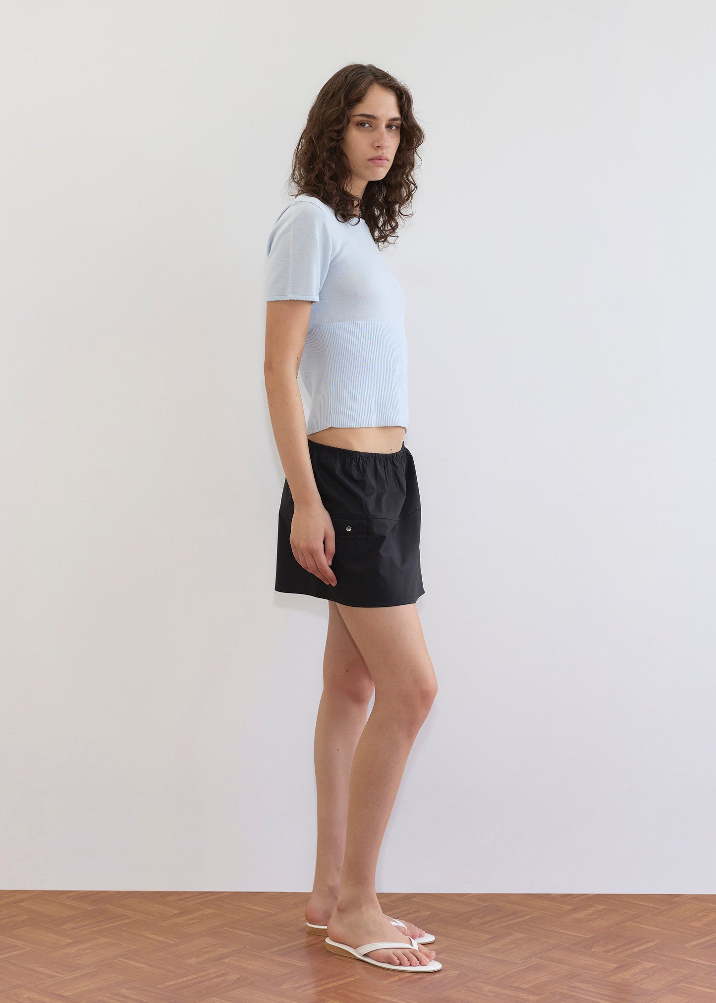 A person with wavy brown hair stands in profile against a plain white wall on a wooden floor, wearing a light blue short-sleeve shirt, white sandals, and the Pocket Skirt - Black.