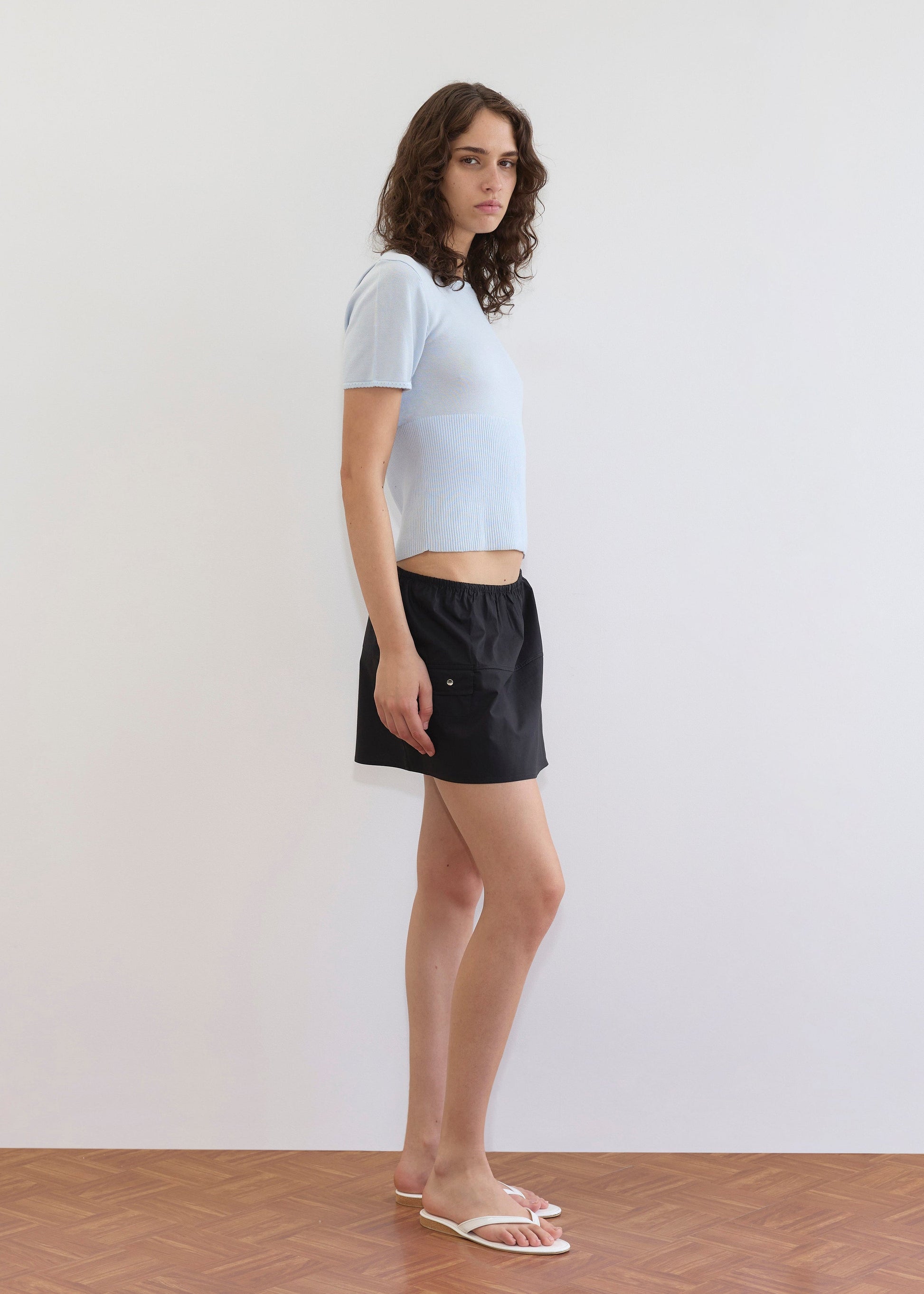 A person with wavy brown hair stands in profile against a plain white wall on a wooden floor, wearing a light blue short-sleeve shirt, white sandals, and the Pocket Skirt - Black.