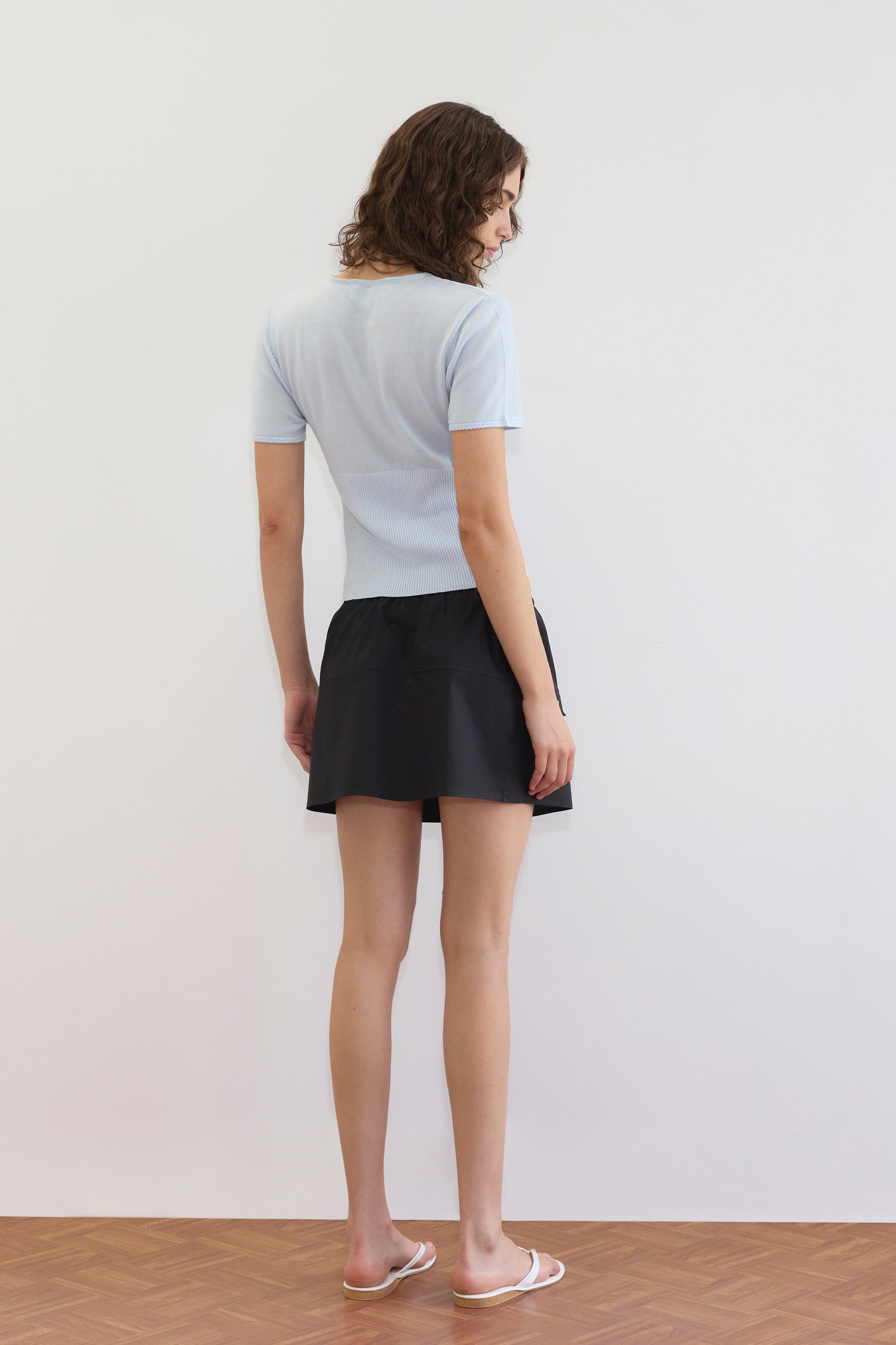 A woman with curly hair faces away, wearing a light blue short-sleeve top, white sandals, and the Pocket Skirt - Black. She stands on a wooden floor against a plain white wall.