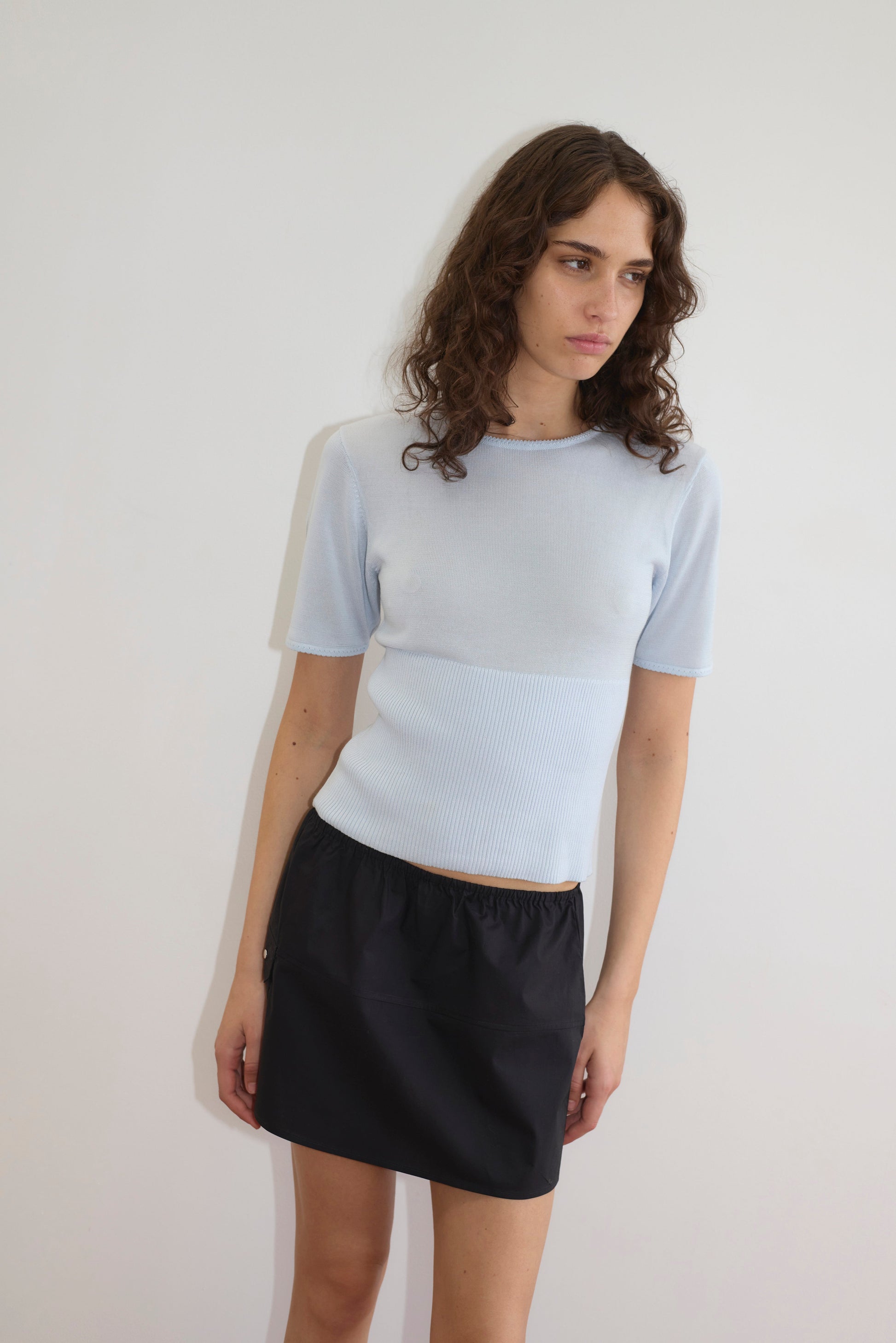A woman with long, wavy brown hair stands against a white wall, wearing a light blue short-sleeve top and the Pocket Skirt - Black. She gazes slightly downward with a neutral expression.