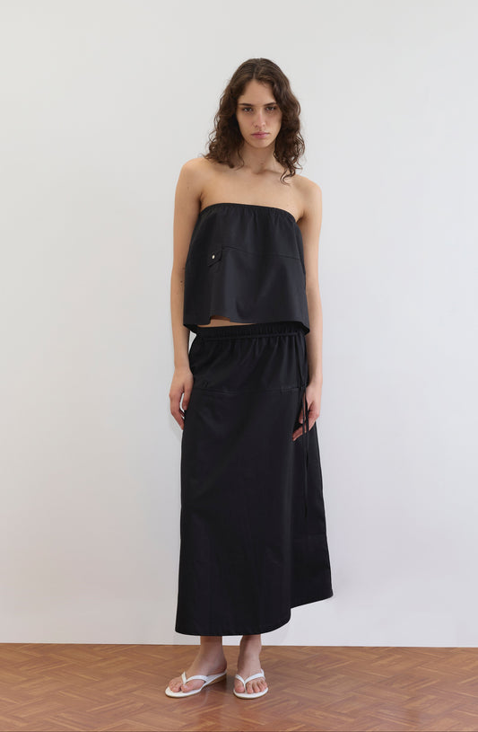 A woman with wavy brown hair stands indoors against a plain white wall, wearing a loose-fitting black strapless top and the Loop Pocket Skirt - Black by Deiji Studios. She pairs the look with white sandals on a wooden floor.