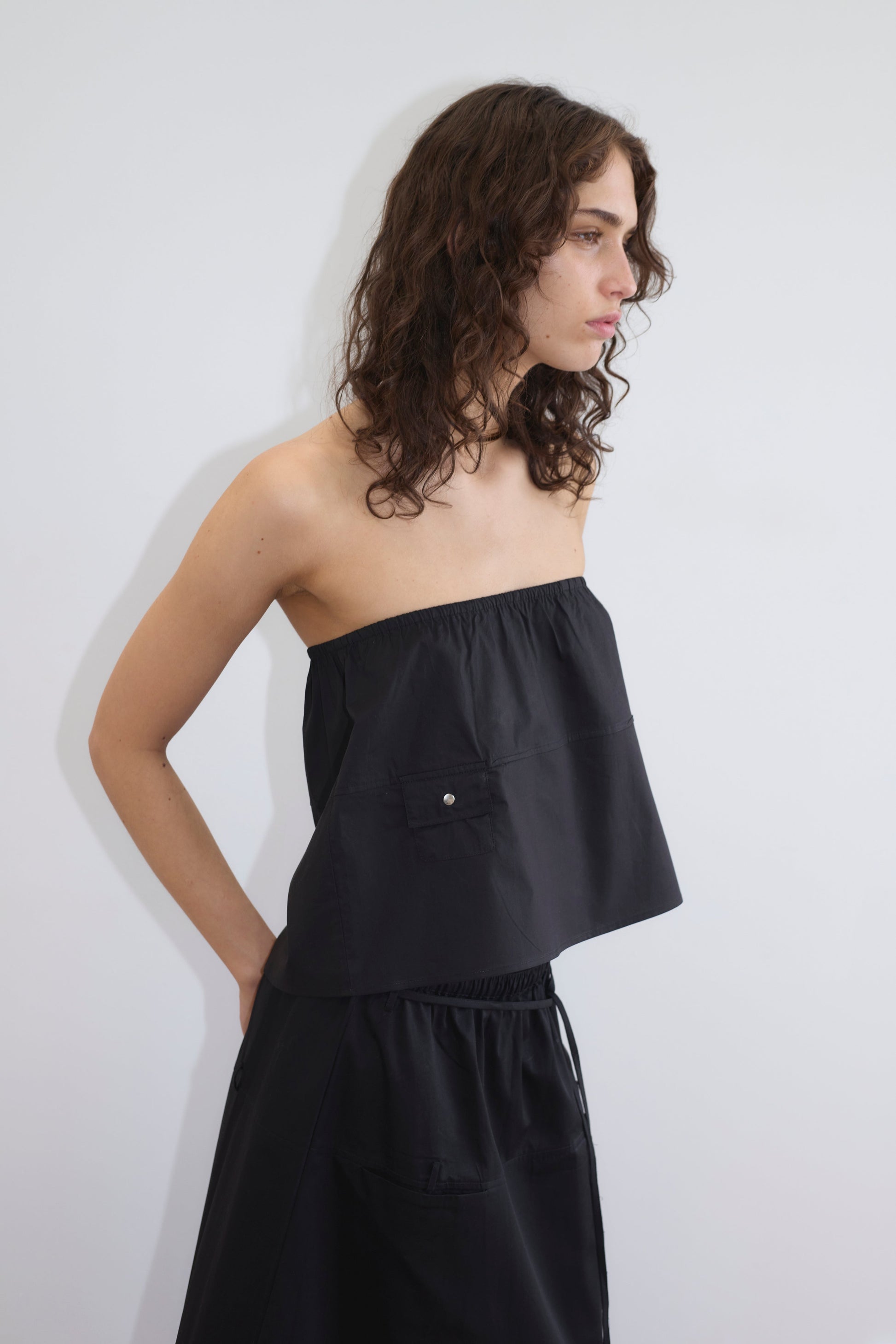 A person with wavy brown hair wears the Pocket Skirt - Black and a strapless black top, standing against a plain white background while looking to the right.