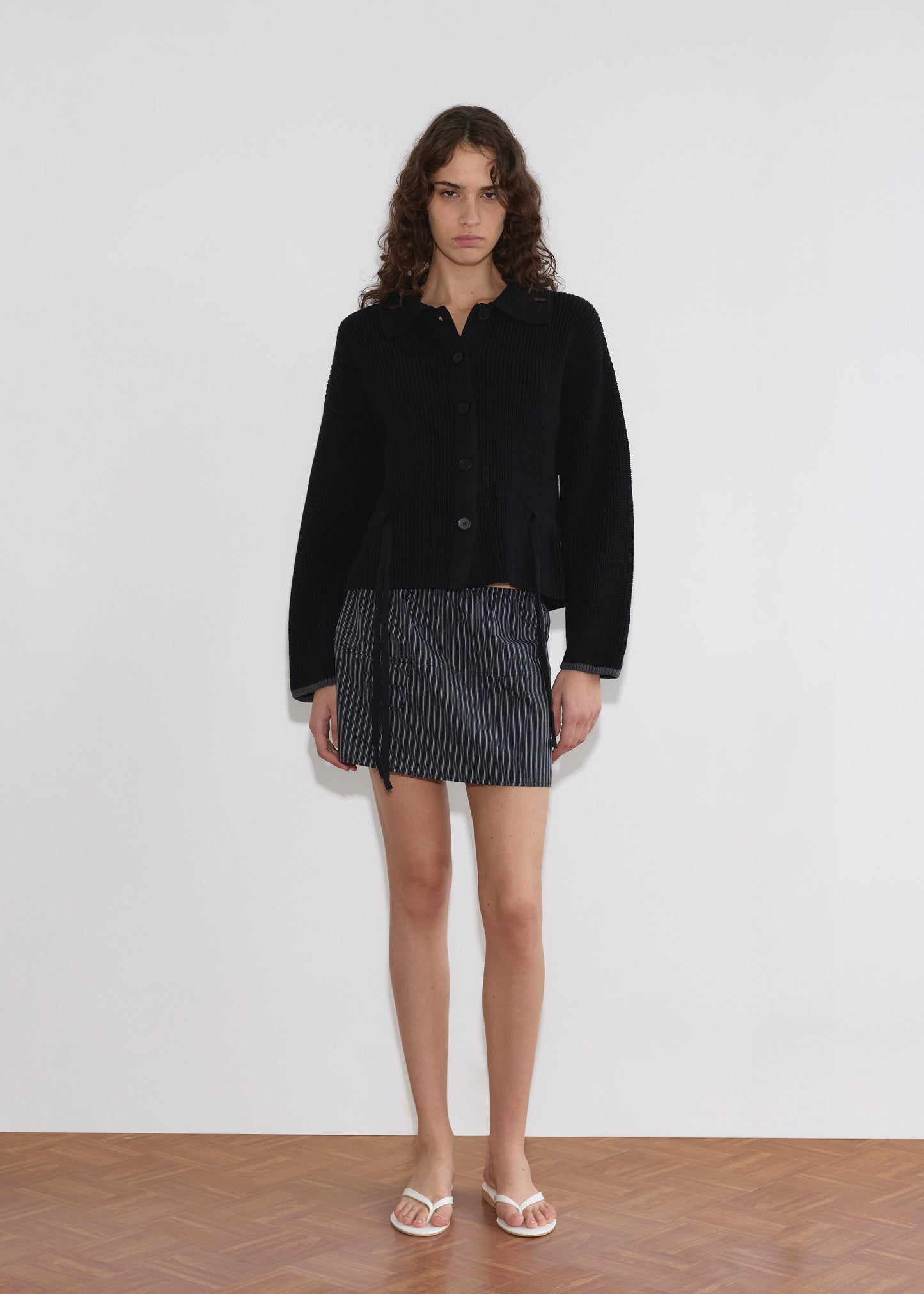 A person with long, curly hair wears the Tie Side Cardigan - Black, a short pinstriped skirt, and white sandals while standing against a plain white wall on a wooden floor.