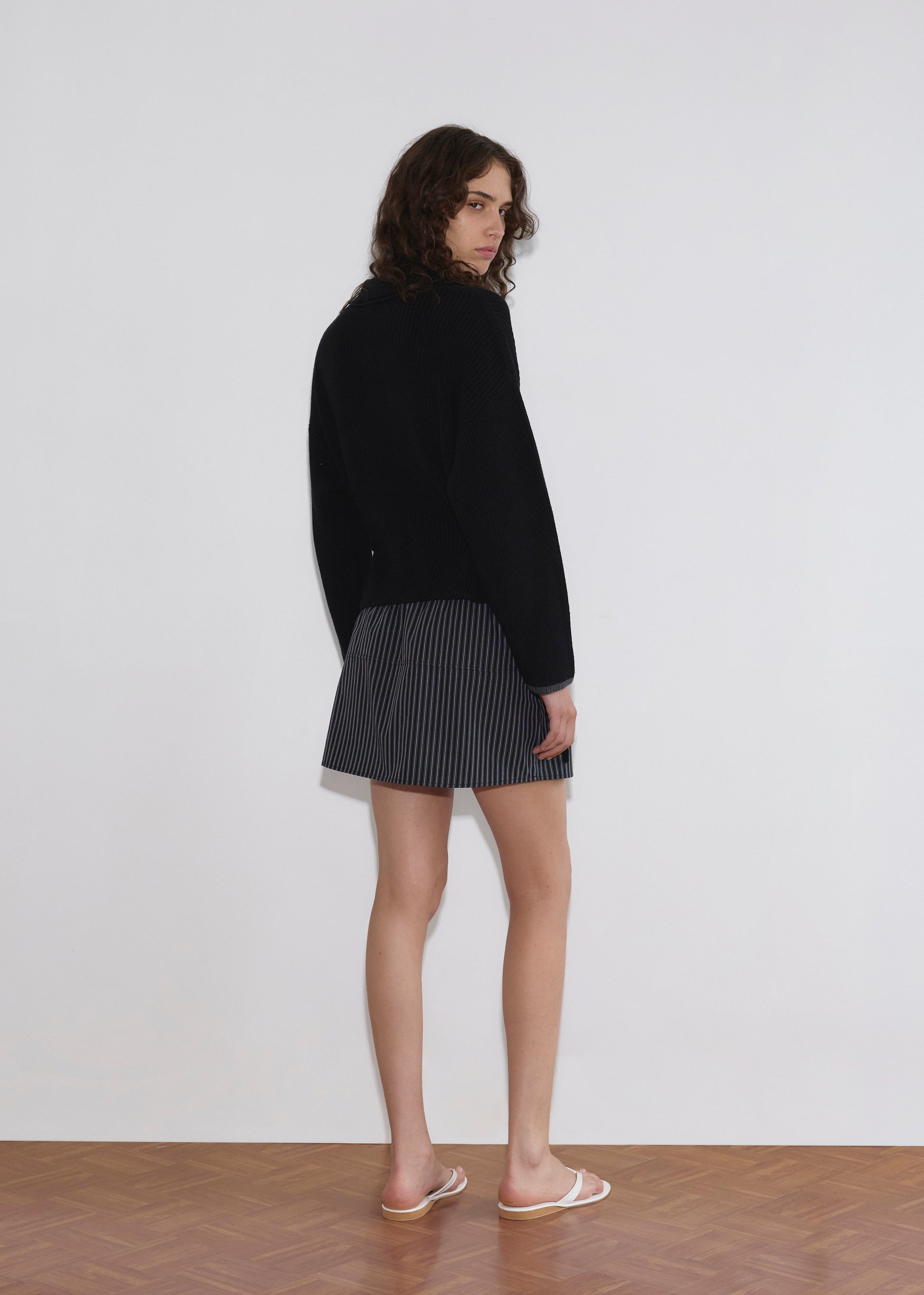 A person with wavy brown hair stands facing away, looking over their shoulder. They wear a Deiji Studios Tie Side Cardigan - Black, a short pinstriped skirt, and white sandals on a wooden floor against a plain white wall.