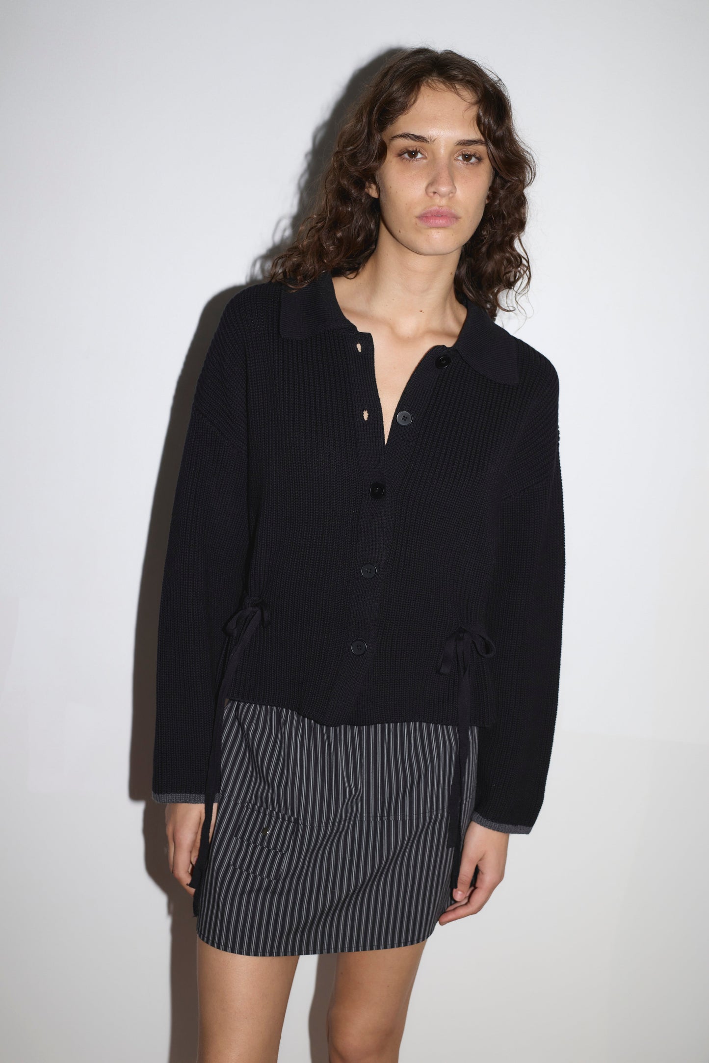 A woman with wavy brown hair stands against a white wall, wearing the Tie Side Cardigan - Black by Deiji Studios and a short, dark skirt with gray stripes. Her relaxed pose reflects the brand’s effortless style.