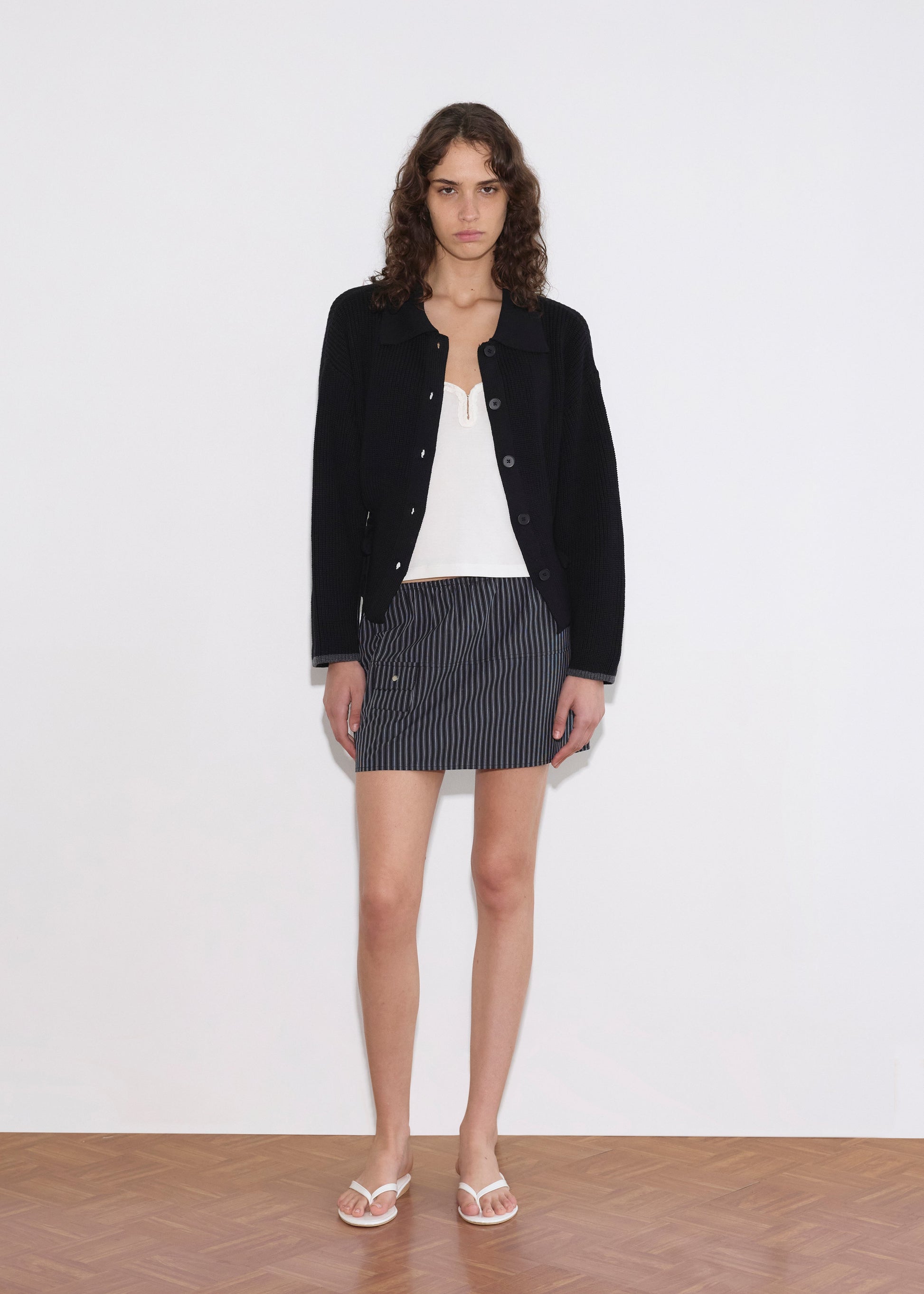 A person with curly hair stands against a plain white wall, wearing the Tie Side Cardigan - Black over a white top, a short pinstriped skirt, and white flip-flops on a wooden floor.