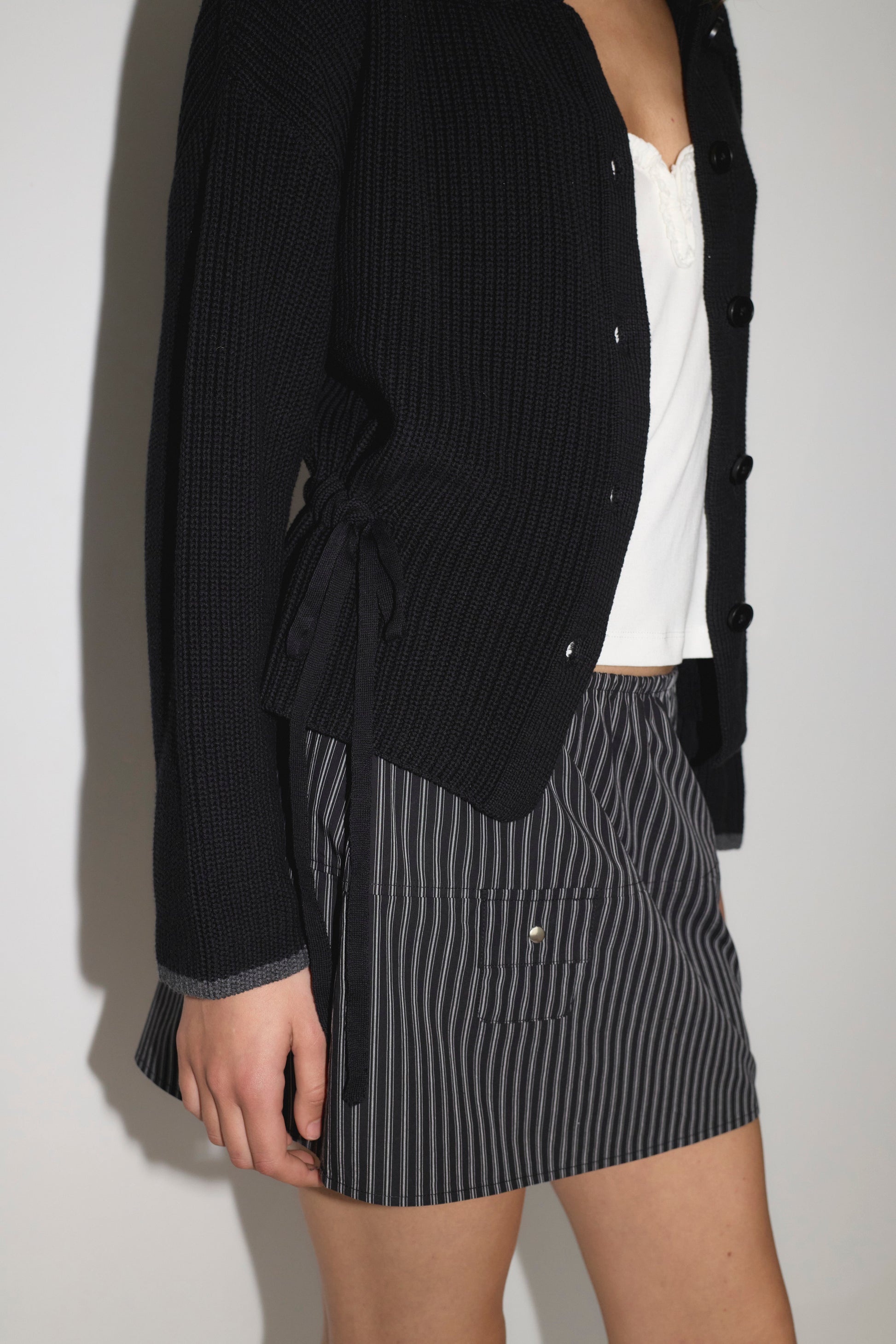 Close-up of a woman wearing the Tie Side Cardigan - Black from Deiji Studios, layered over a white top and paired with a dark pinstripe mini skirt.