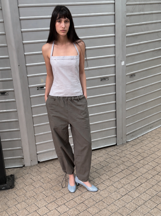 A woman with dark hair and a fringe stands against metal shutters wearing the Hem Toggle Pant - Moss Stripe by Deiji Studios, moss-green trousers with a toggle-gathered hem, with a pale striped halter top and pale blue heels.