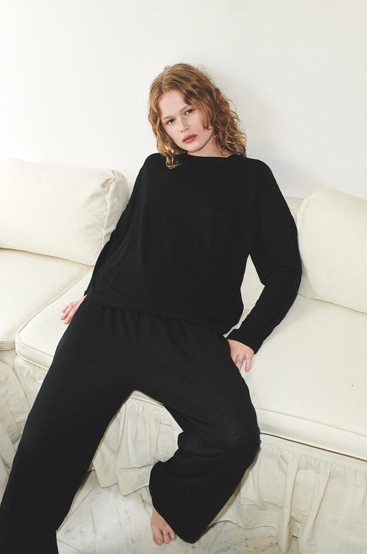 A relaxed woman with wavy, shoulder-length hair sits barefoot on a white couch, wearing the Soft Long Sleeve Top in black with matching pants and gazing toward the camera.
