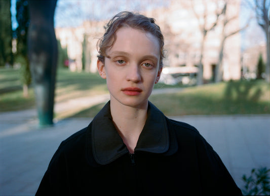 A young person with light skin and blond hair wears the Collared Zip Jacket - Black Contrast, standing outdoors on a paved path with blurred green grass and trees in the background.