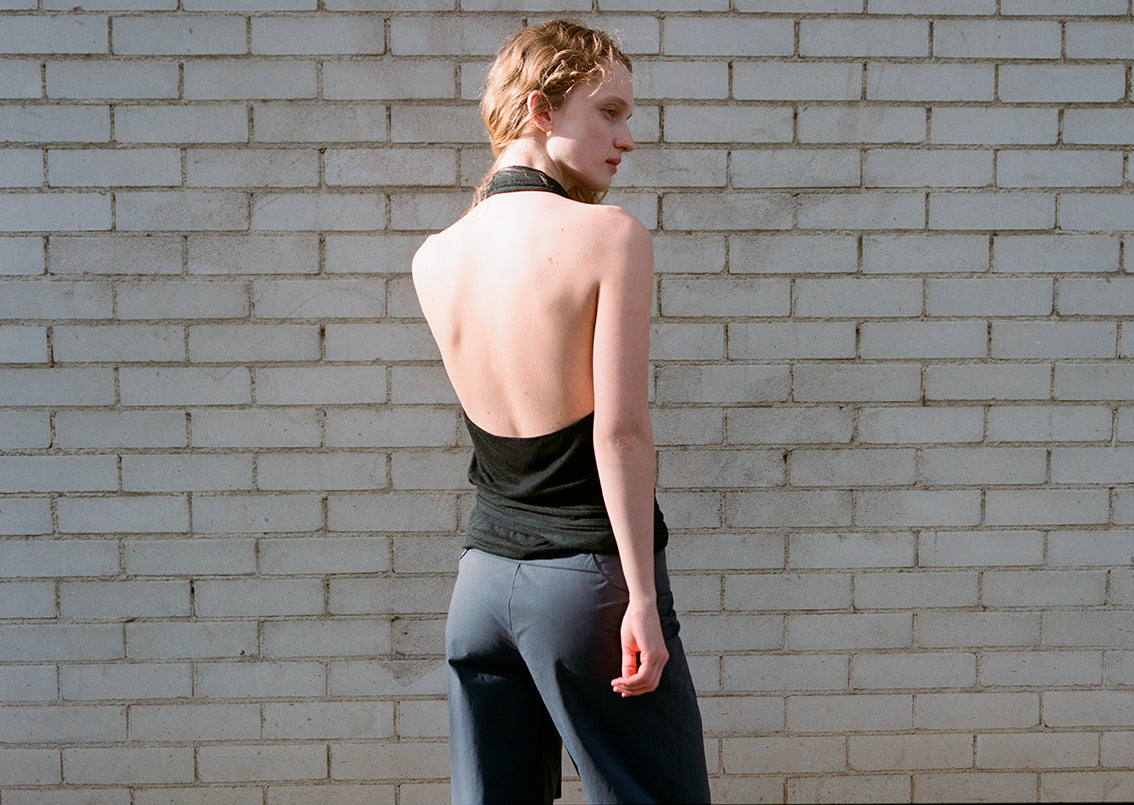 A person with short, light hair stands in front of a white brick wall, turned away from the camera. They wear a black backless halter top and Deiji Studios' Panelled Pant - Charcoal with a straight-leg silhouette, looking over their shoulder.