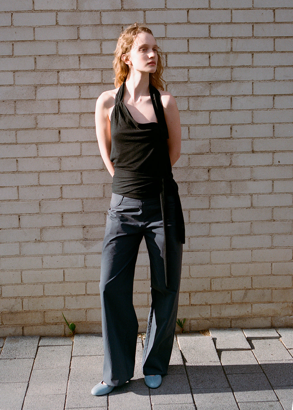 A woman stands outside by a light brick wall, wearing a black halter top, Deiji Studios Panelled Pant - Charcoal with a straight-leg silhouette, and light blue shoes. Her hands are behind her back as she looks to the side.