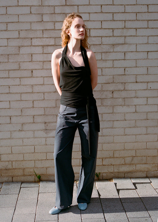 A woman stands outside by a light brick wall, wearing a black halter top, Deiji Studios Panelled Pant - Charcoal with a straight-leg silhouette, and light blue shoes. Her hands are behind her back as she looks to the side.