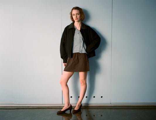 A person stands indoors by a plain light blue wall, wearing a black jacket, gray shirt, Deiji Studios Pocket Skirt - Sepia, and black flats. They have short wavy hair and stand relaxed with one hand on their hip.