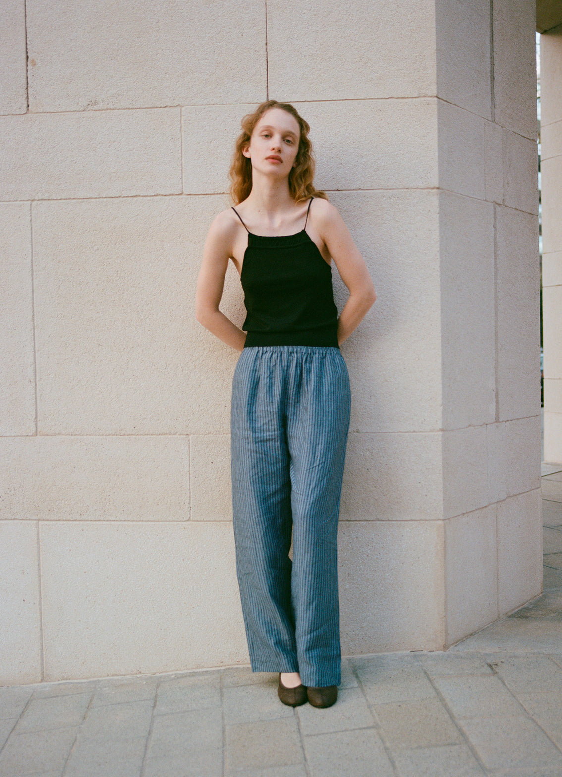 A woman stands outdoors against a light stone wall, wearing the Drawcord Knit Top - Black by Deiji Studios with loose gray striped pants and dark shoes. Her hands are behind her back as she gazes calmly at the camera.
