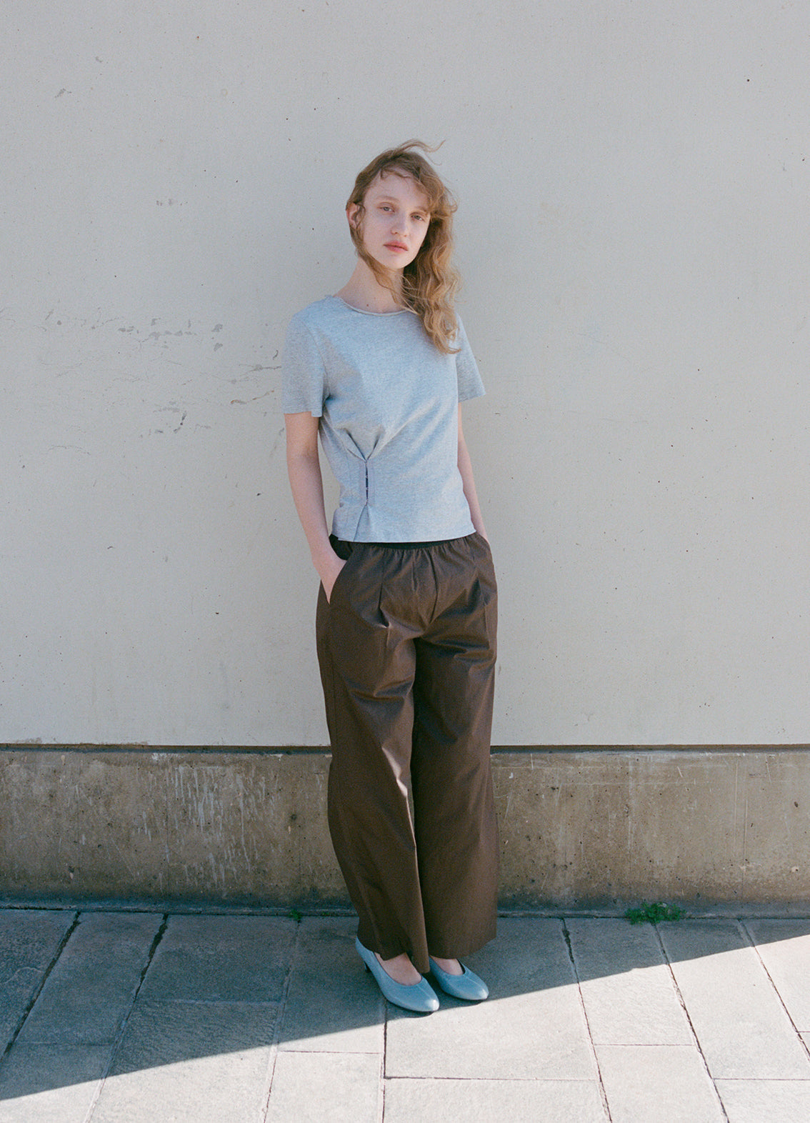 A woman stands against a plain light wall, wearing the Pinch Tee - Grey from Deiji Studios and loose brown pants. She pairs her outfit with light blue shoes, has one hand in her pocket, and gazes neutrally at the camera.