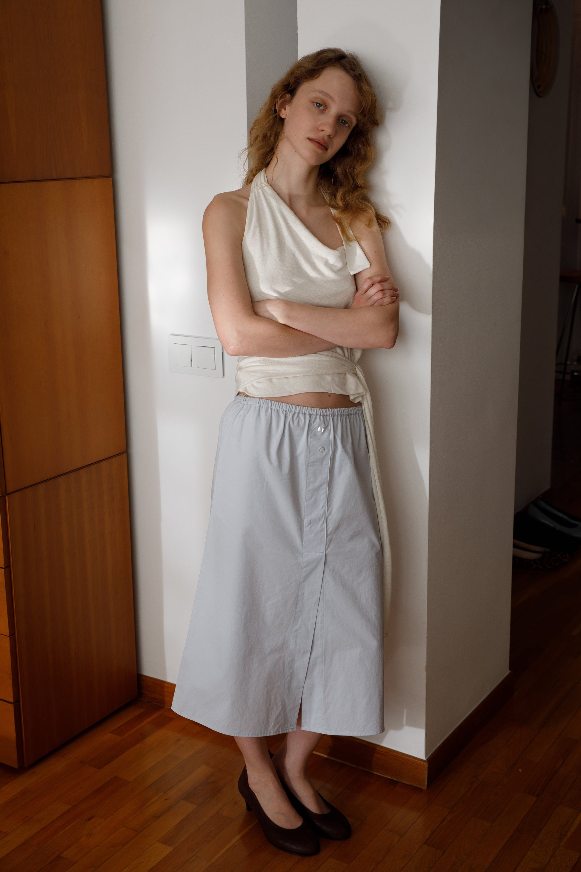 A woman with wavy blonde hair, wearing a sleeveless white top and the Overlap Skirt - Blue by Deiji Studios, stands indoors against a white wall with arms crossed, gazing calmly at the camera.