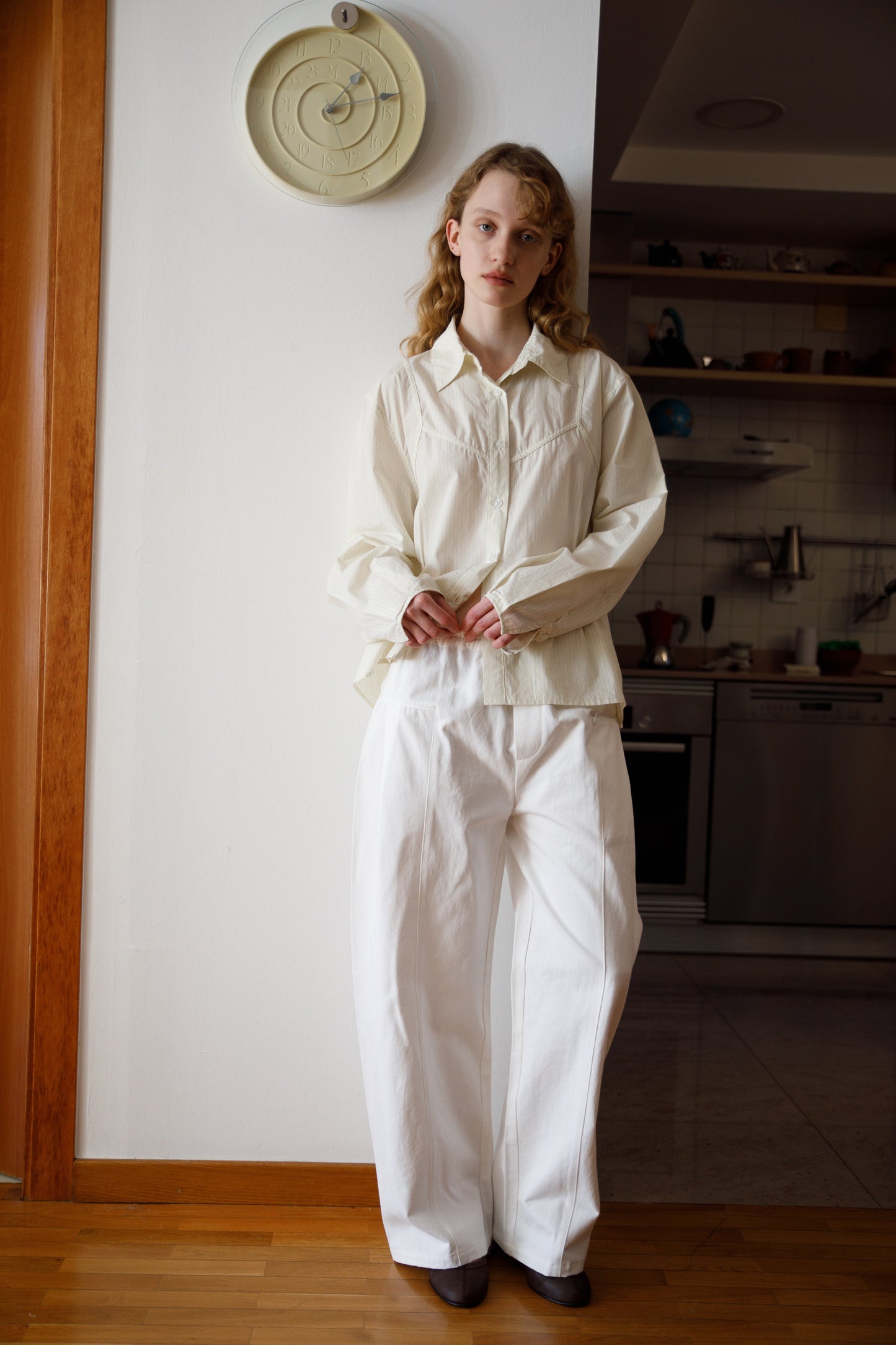 A young woman with long hair stands indoors near a kitchen, wearing a loose-fitting cream blouse and the Pleat Pant - White. She is by a white wall with a round clock above her, standing on wooden flooring.
