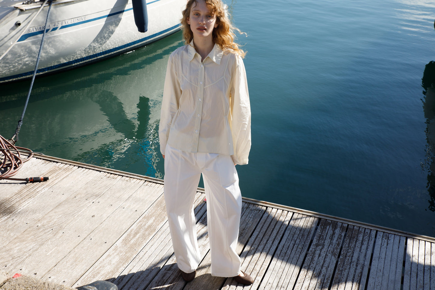 A woman with wavy red hair stands on a wooden dock wearing a cream button-up shirt and the Pleat Pant - White. A sailboat and calm blue-green water are visible in the background.