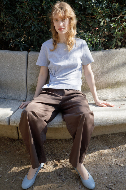 A person with wavy, light hair sits on a stone bench outdoors, wearing a grey Pinch Tee by Deiji Studios, loose brown pants, and light gray shoes. Bushes and sunlight appear in the background.